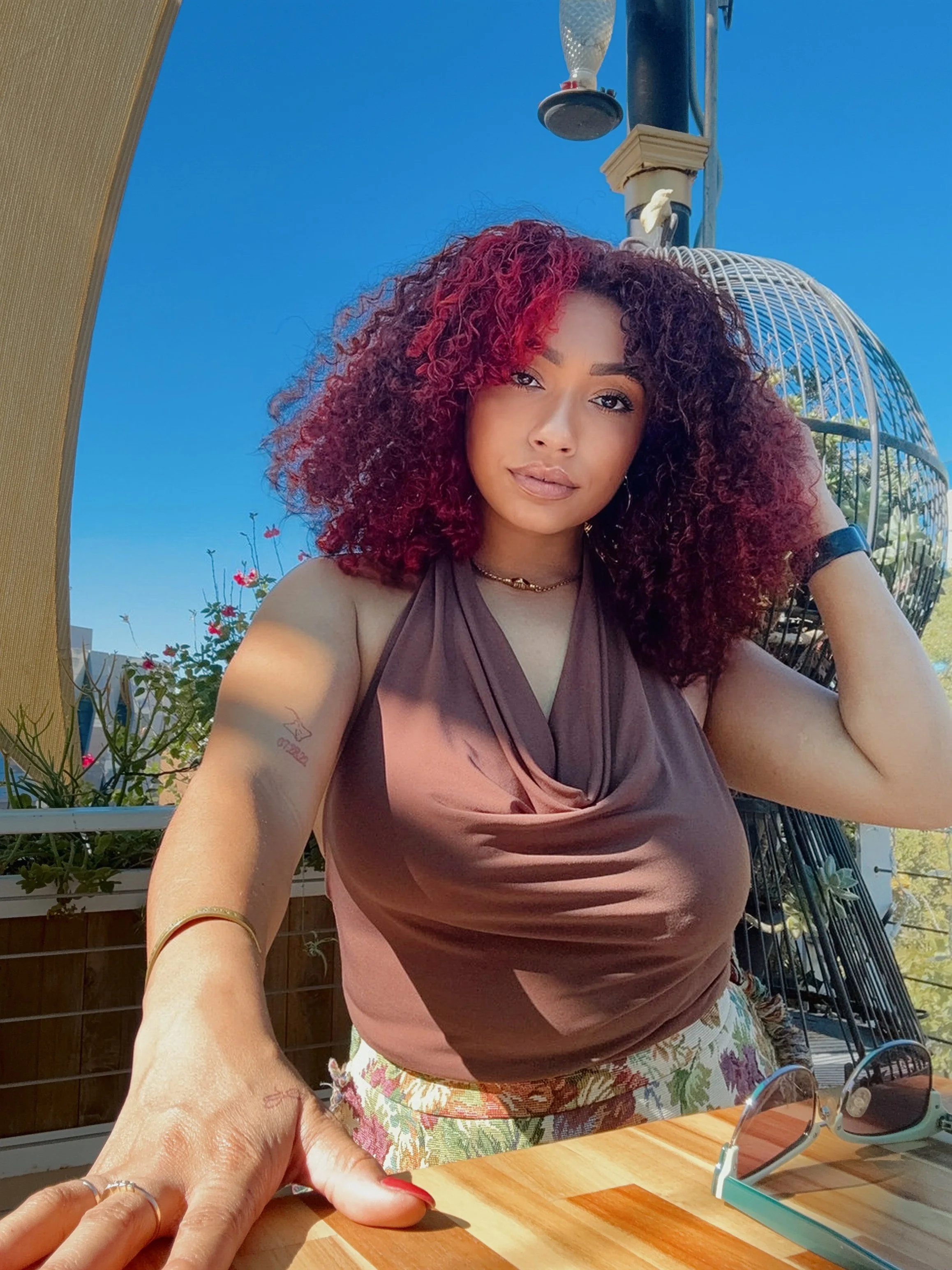 A woman with curly, burgundy and dark hair, wearing a brown sleeveless top and floral pants, poses outdoors against a blue sky with a birdcage and plants in the background.