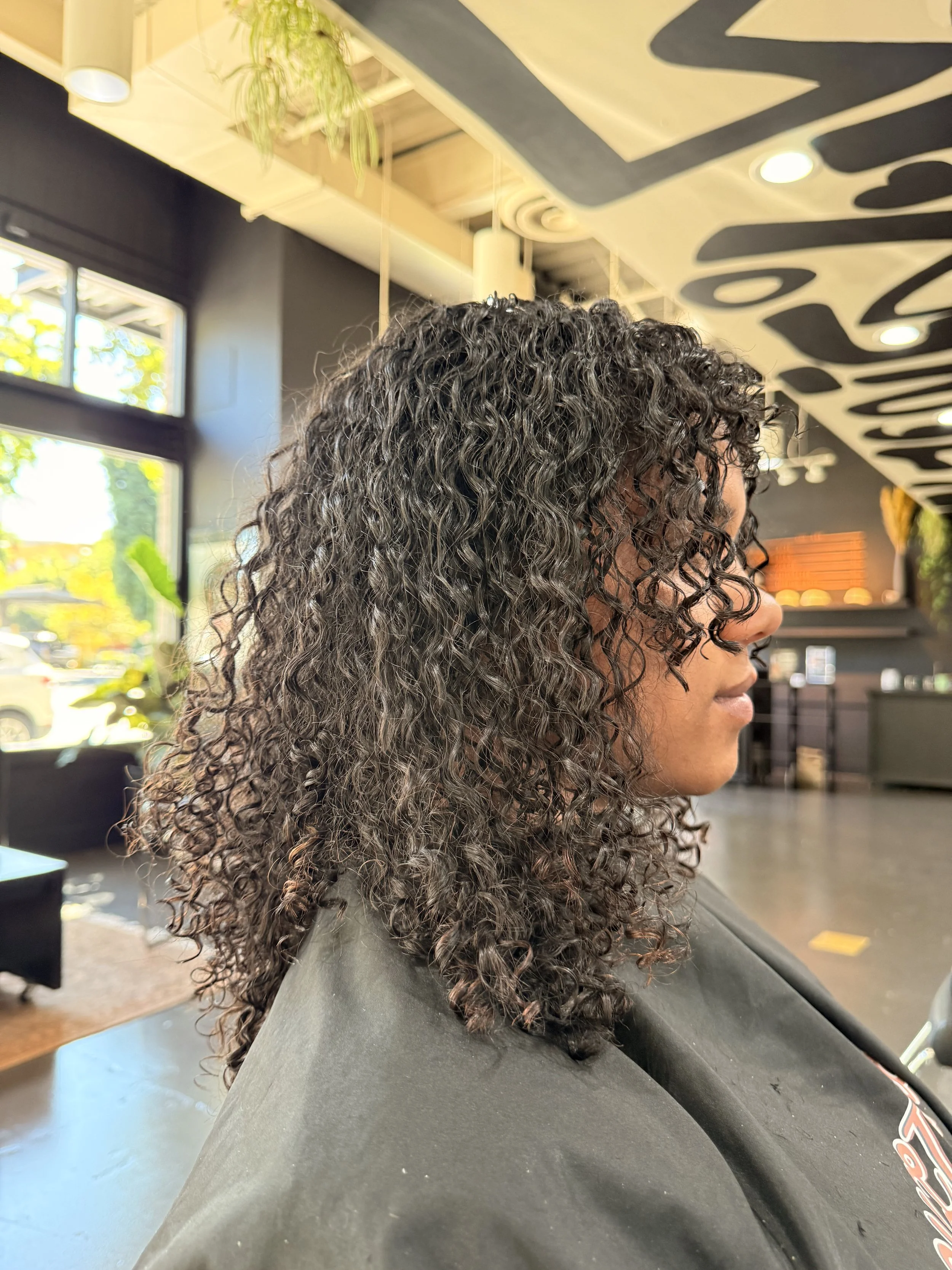 Profile of a woman with dark, curly hair sitting in a salon.
