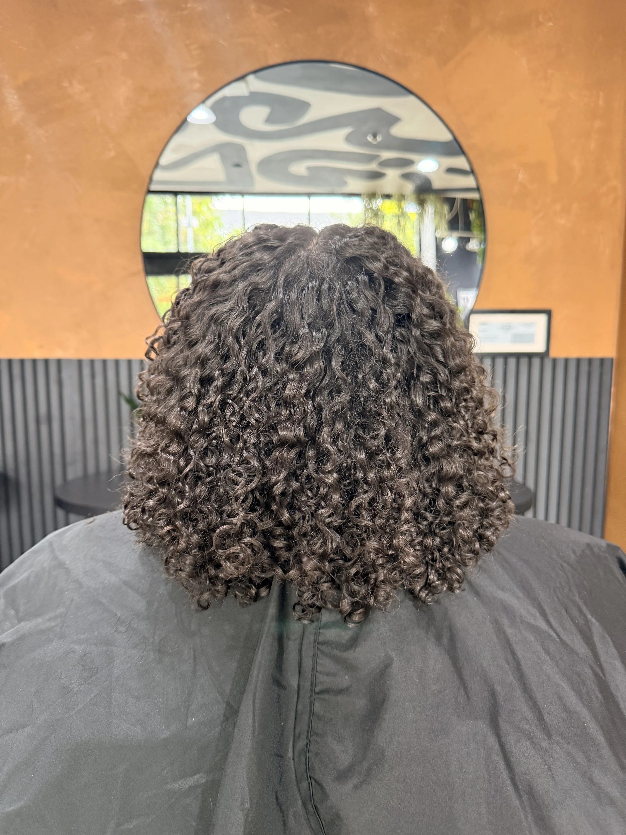 Back of a person with curly, shoulder-length hair sitting in a salon chair, seen through a mirror.