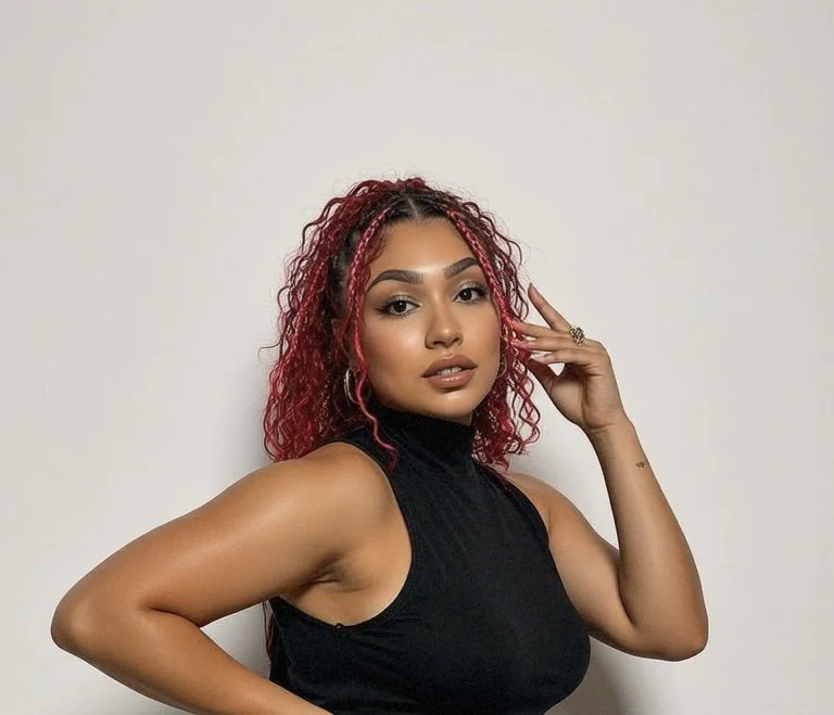 A woman with red curly hair wearing a sleeveless black top, standing in front of a plain white wall.