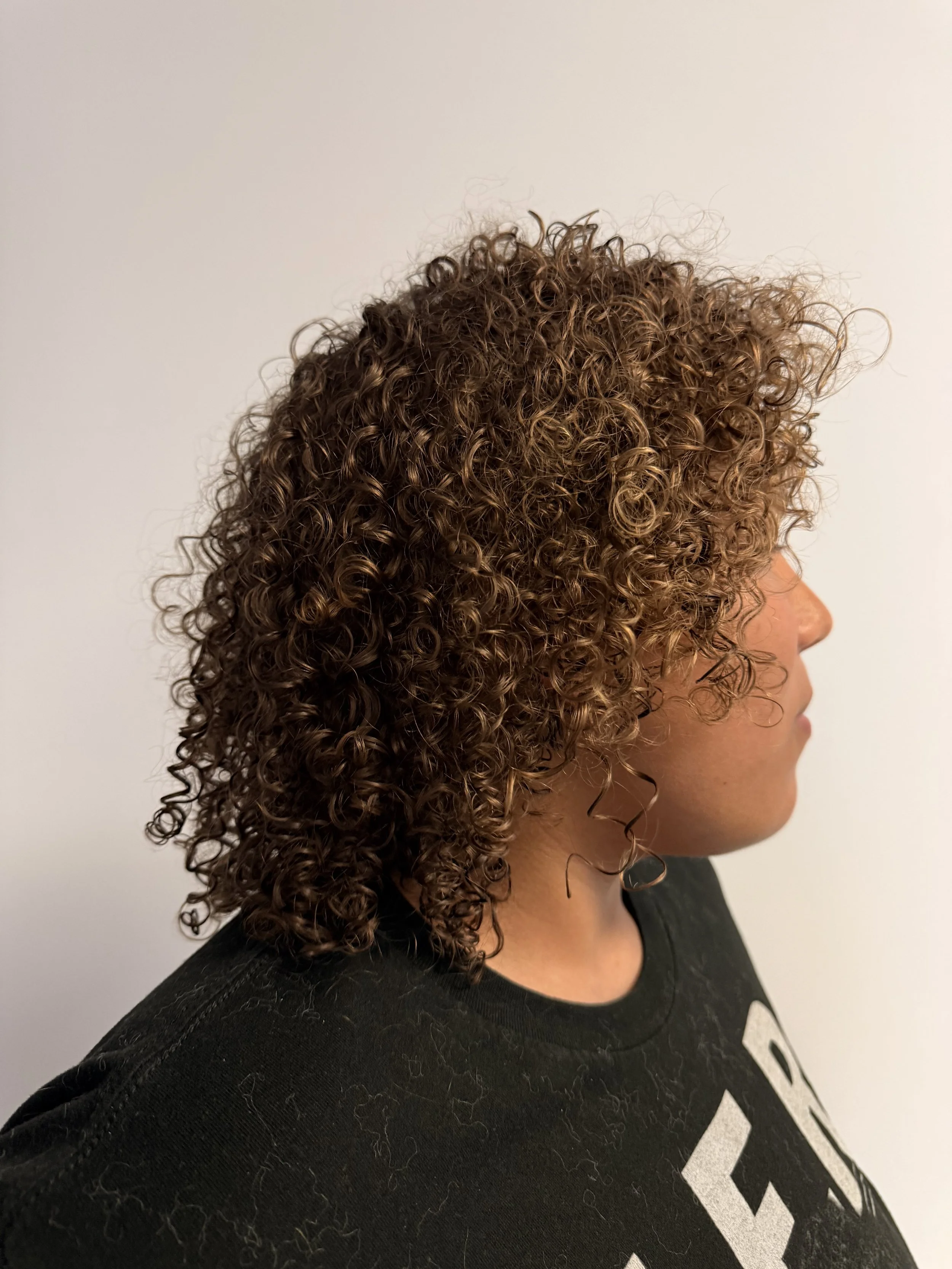Side profile of a woman with light brown, curly hair, wearing a black T-shirt, against a plain white background.