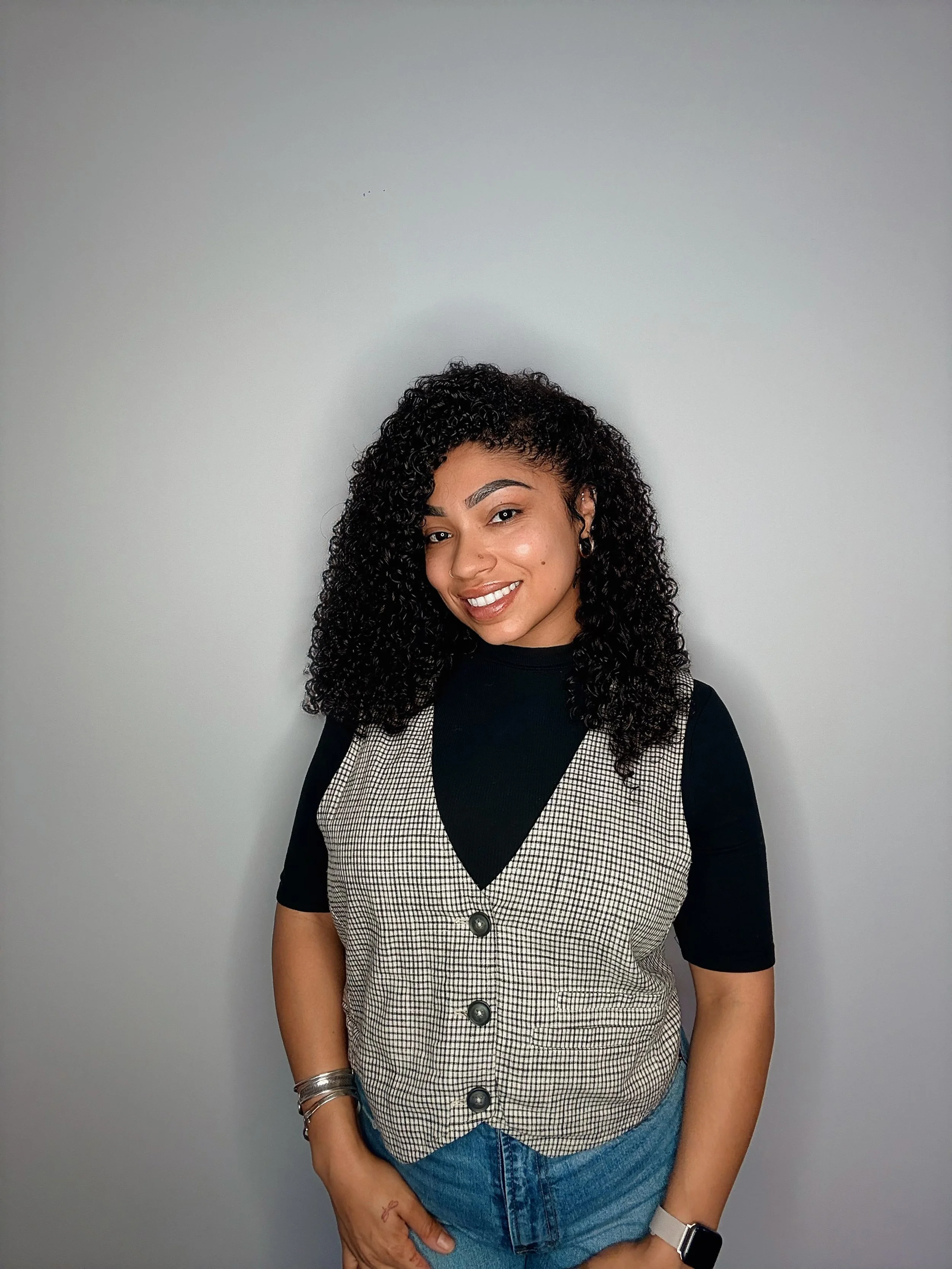 A smiling woman with curly black hair wearing a black shirt, a beige checkered vest, blue shorts, and accessories including earrings, bracelets, a ring, and a smartwatch, standing against a plain gray background.