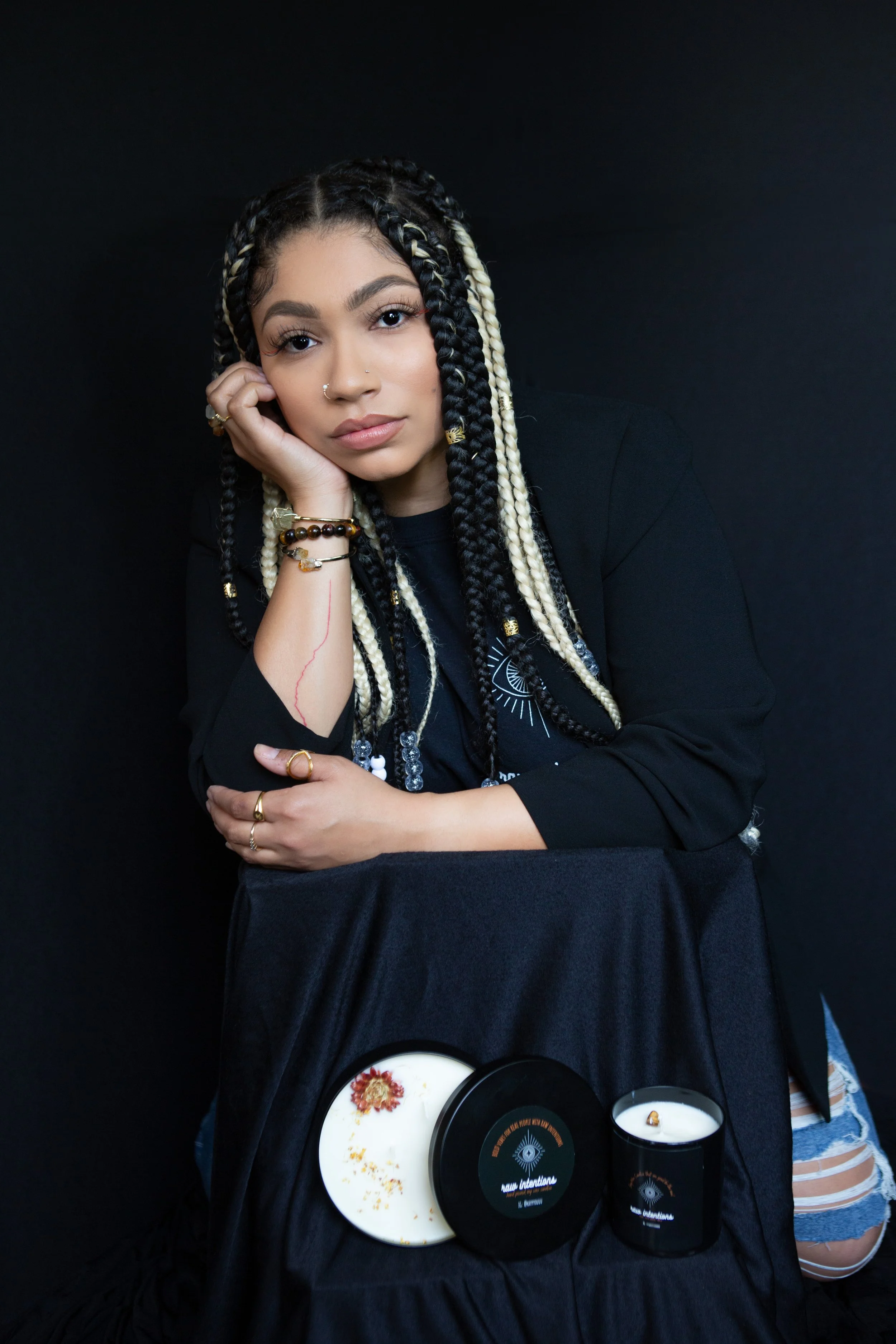 A young woman with braided hair and jewelry resting her head on her hand, sitting at a table with a black background. She has tattoos and is dressed in black. On the table in front of her are black containers with white labels, a white plate with a floral design, and a small candle.
