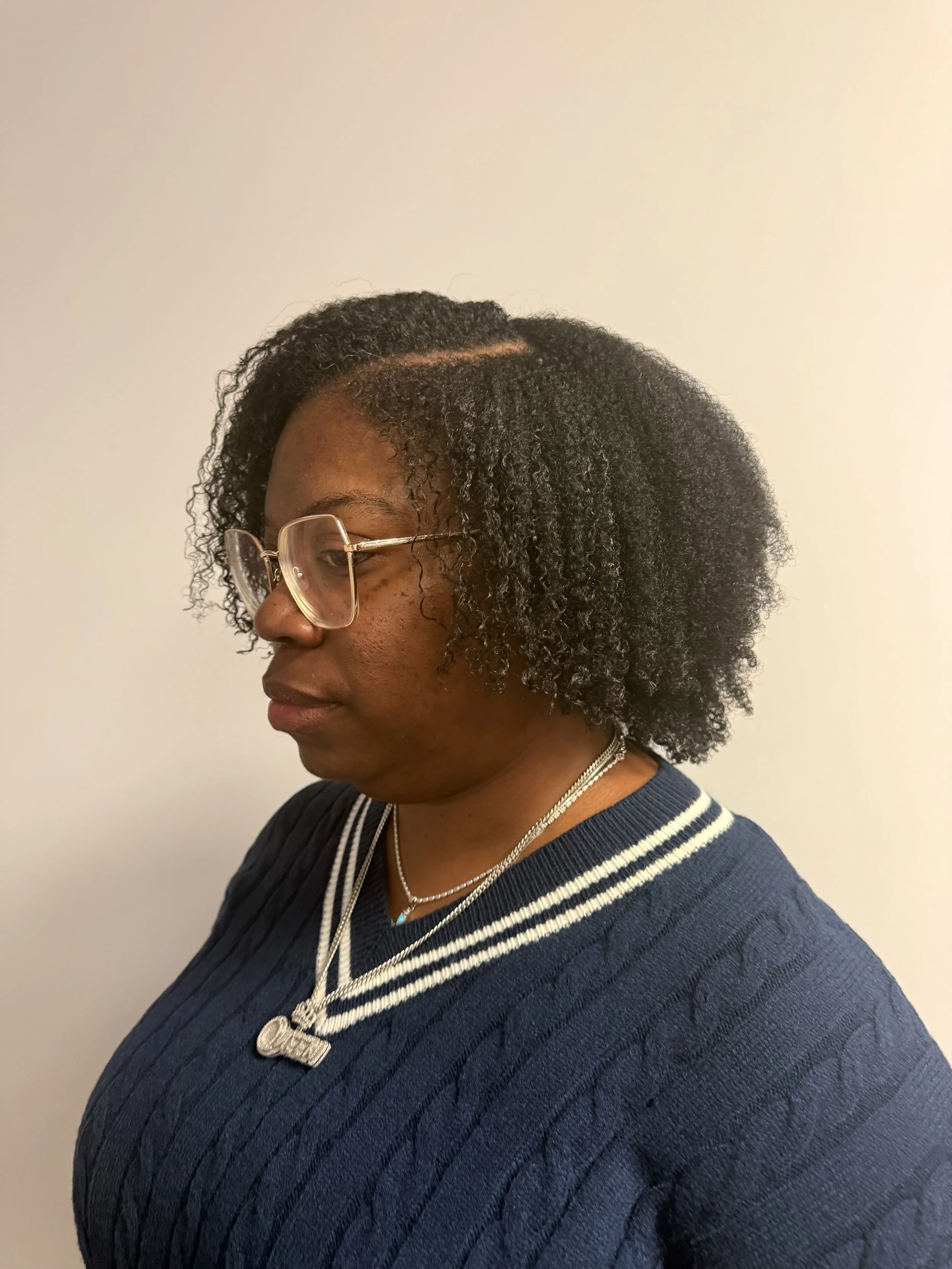 A woman with dark, curly hair wearing glasses and a navy blue sweater with white stripes on the collar, standing against a plain light-colored wall.
