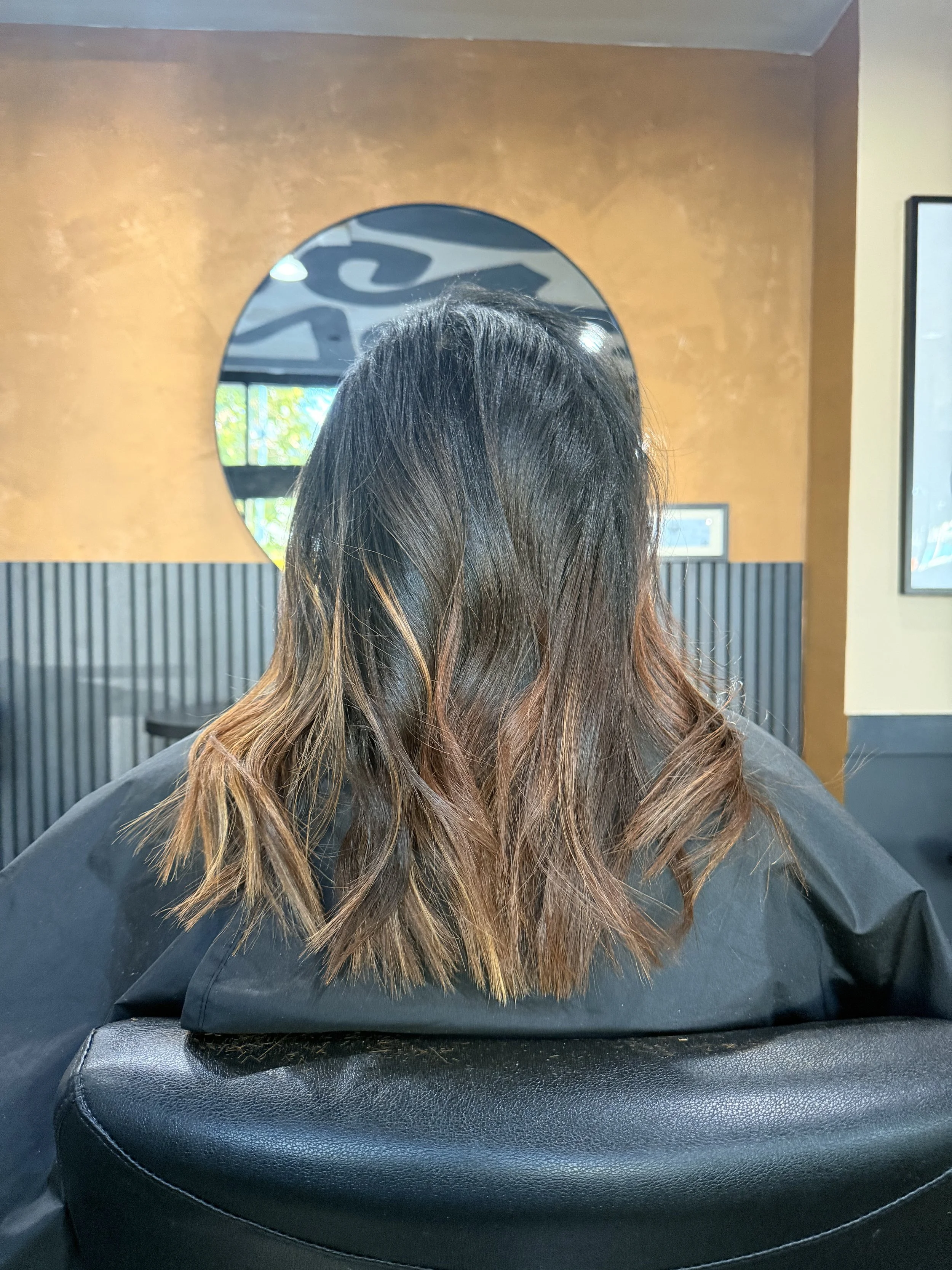 Back view of a woman seated at a salon, showing her shoulder-length hair with dark roots and lighter brown, slightly wavy ends, in a hair salon with a mirror and decorative wall.