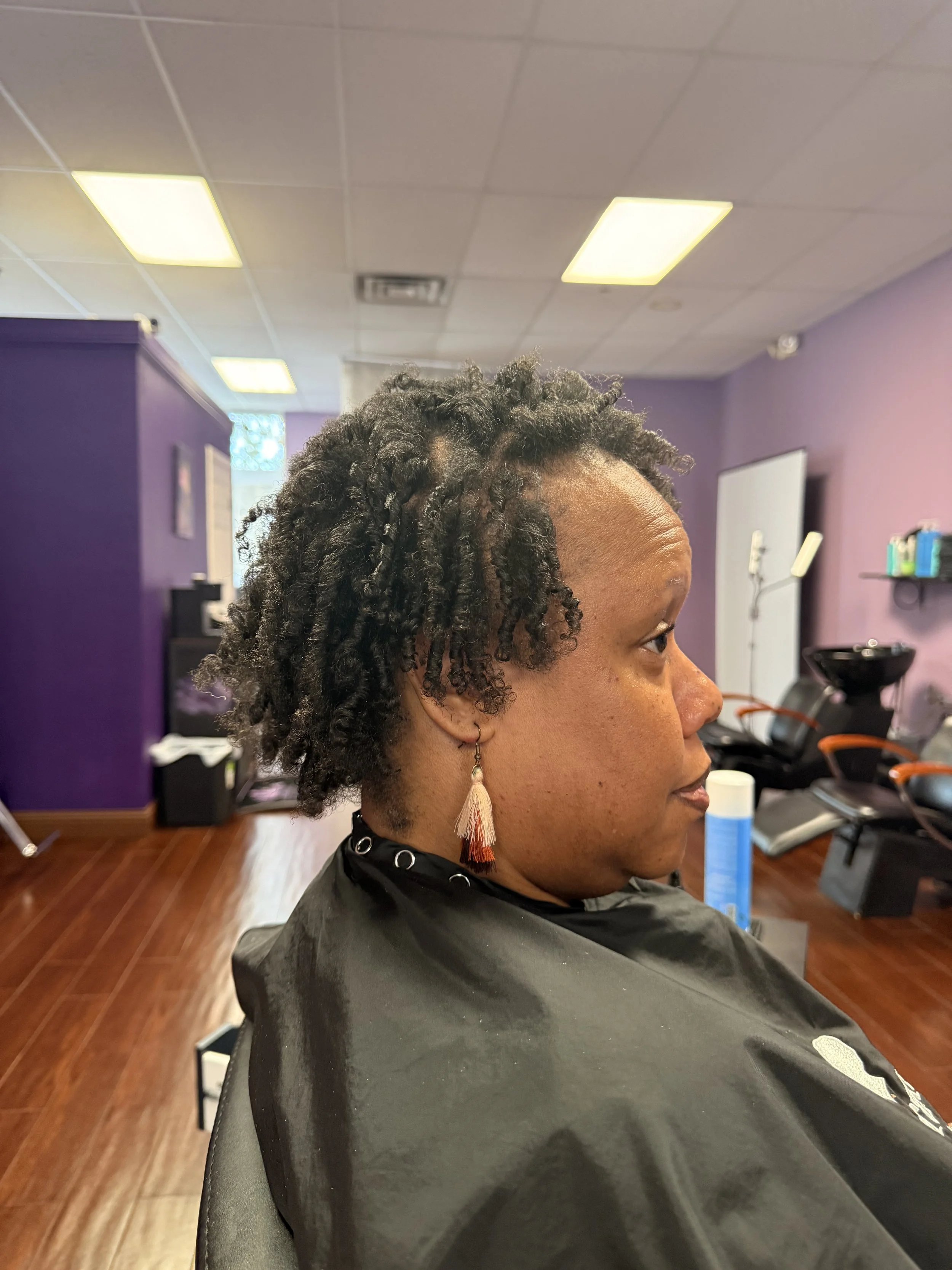 Profile of a woman with short, curly, natural hair sitting in a salon chair, wearing a black cape, in a salon with purple walls.