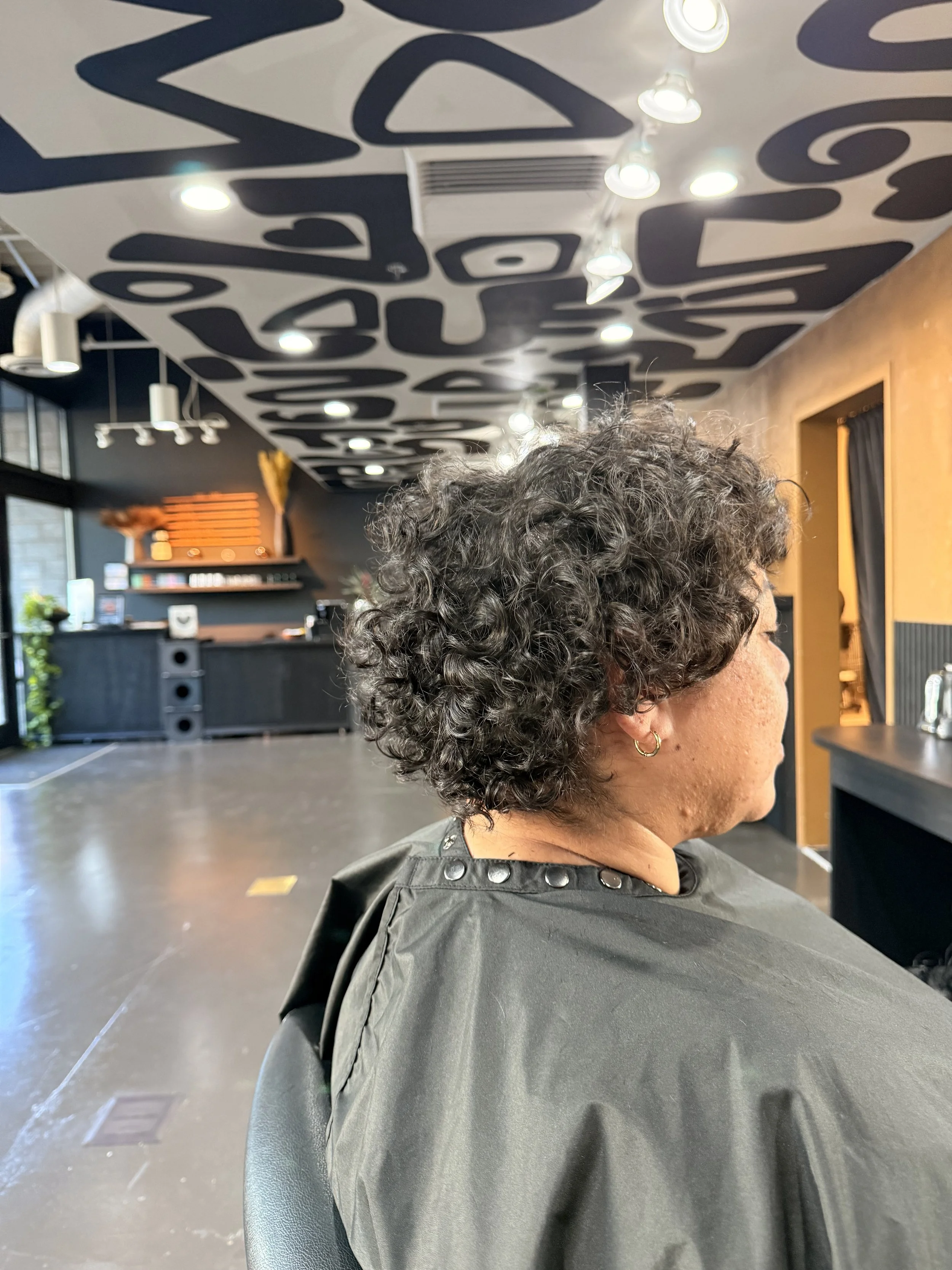 A woman with curly short hair sitting in a hair salon chair, with a black salon cape on, inside a modern hair salon with black and brown interior design and decorative ceiling artwork.
