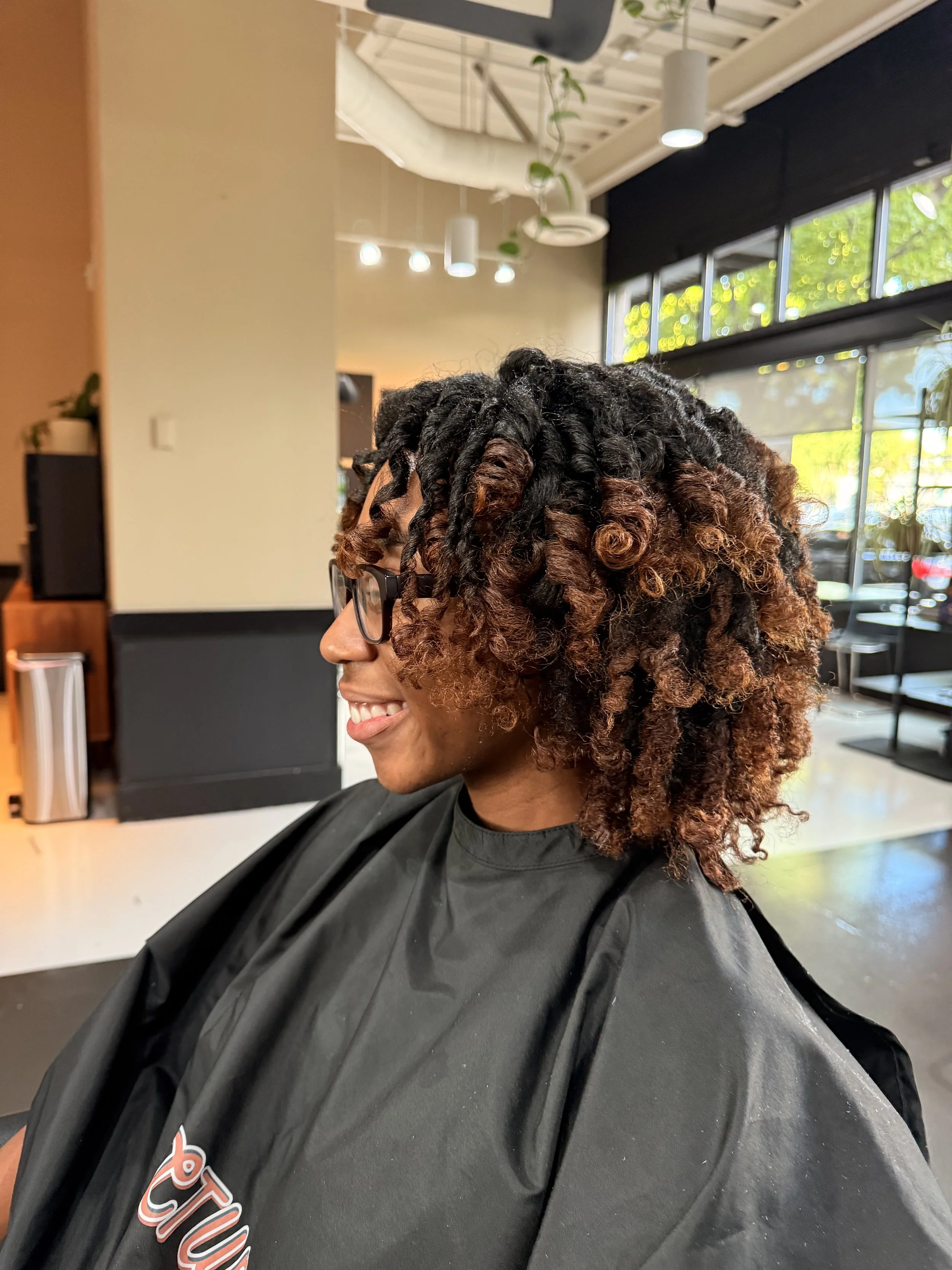 A woman with glasses and curly two-tone hair, from dark brown to a reddish-brown, smiling while sitting in a hair salon.
