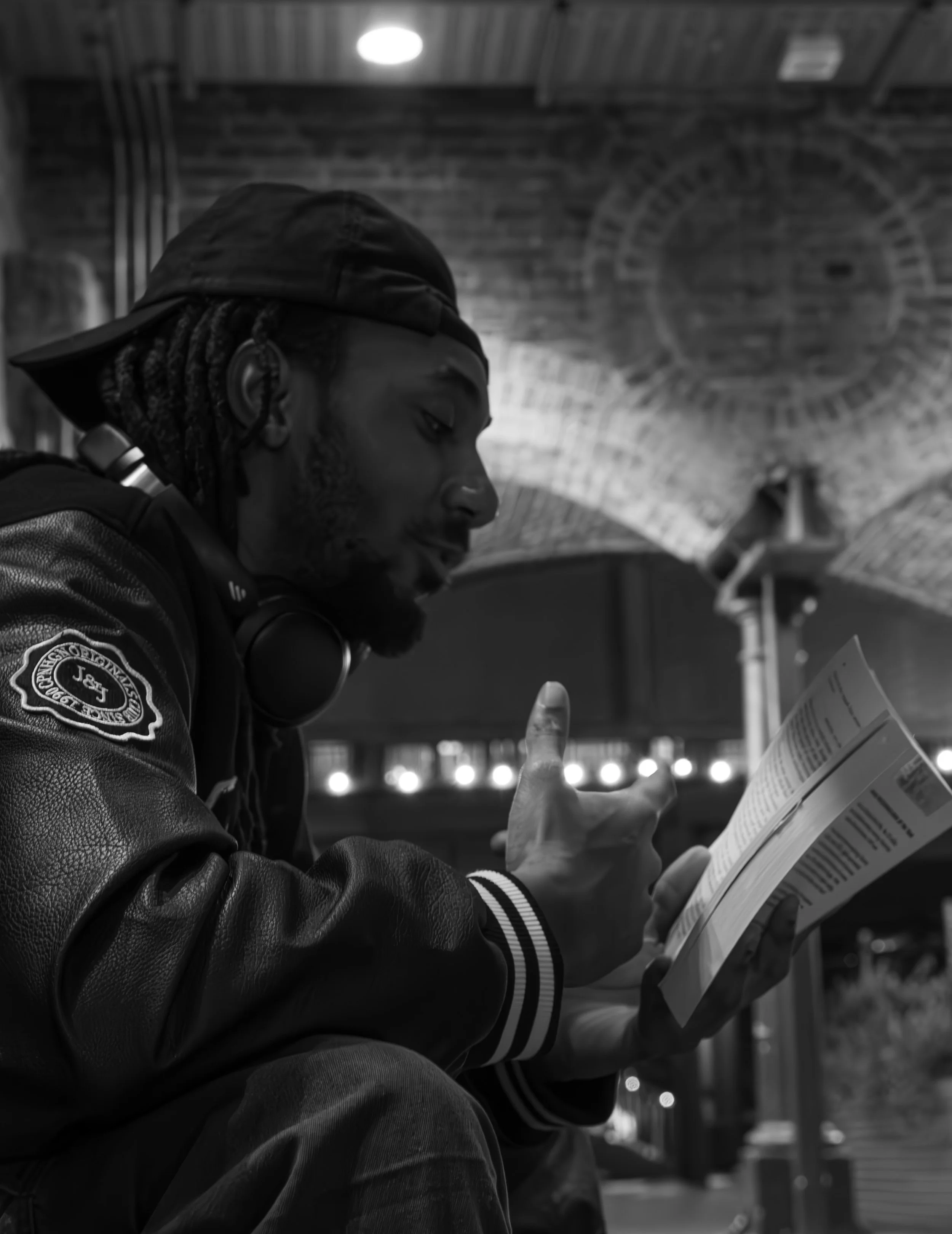 A man with dreadlocks wearing a baseball cap and headphones around his neck, reading a pamphlet or brochure in a setting with a brick ceiling and string lights.