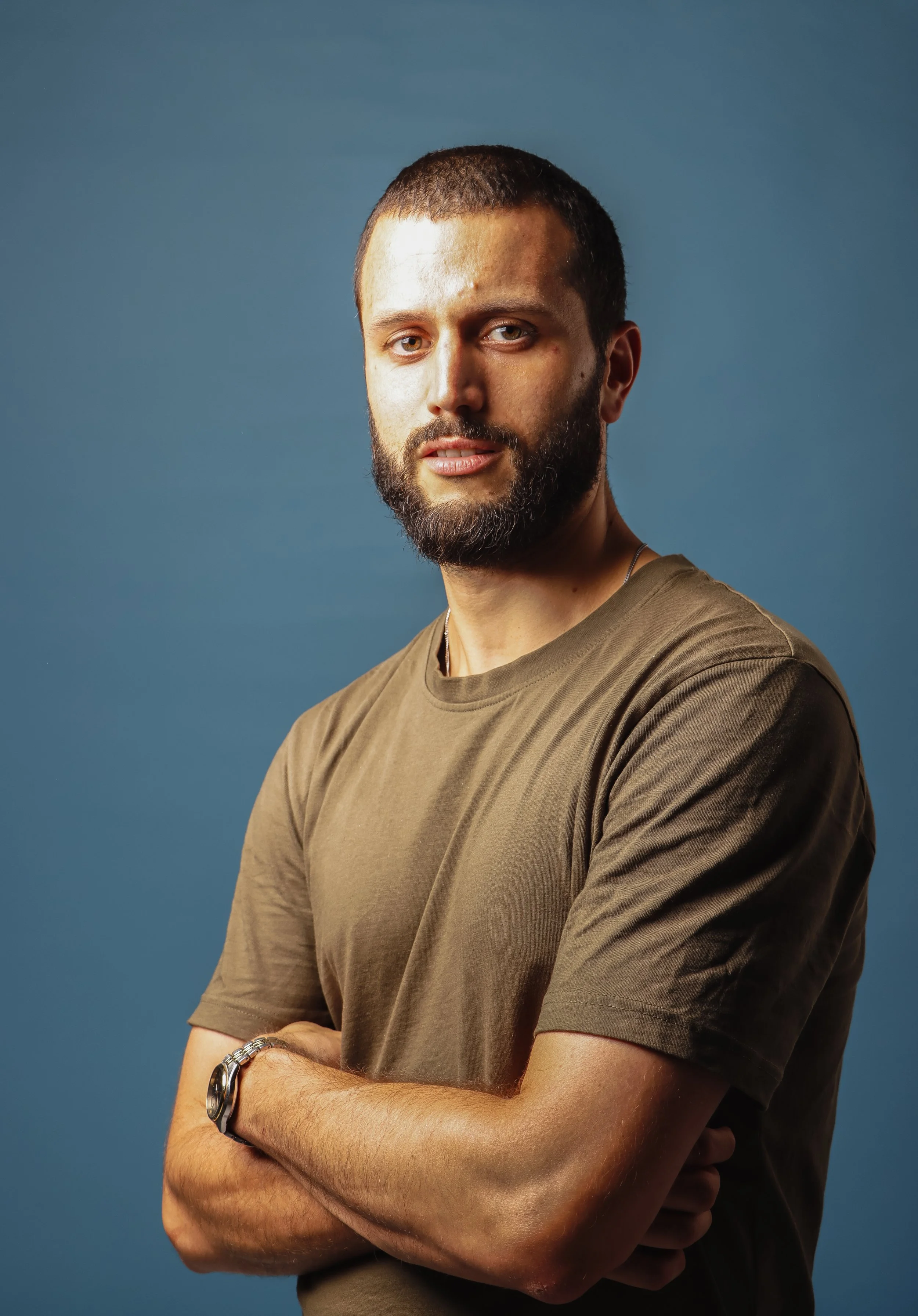 A young man with a beard and short hair, wearing a brown t-shirt and a wristwatch, standing with arms crossed against a solid blue background.