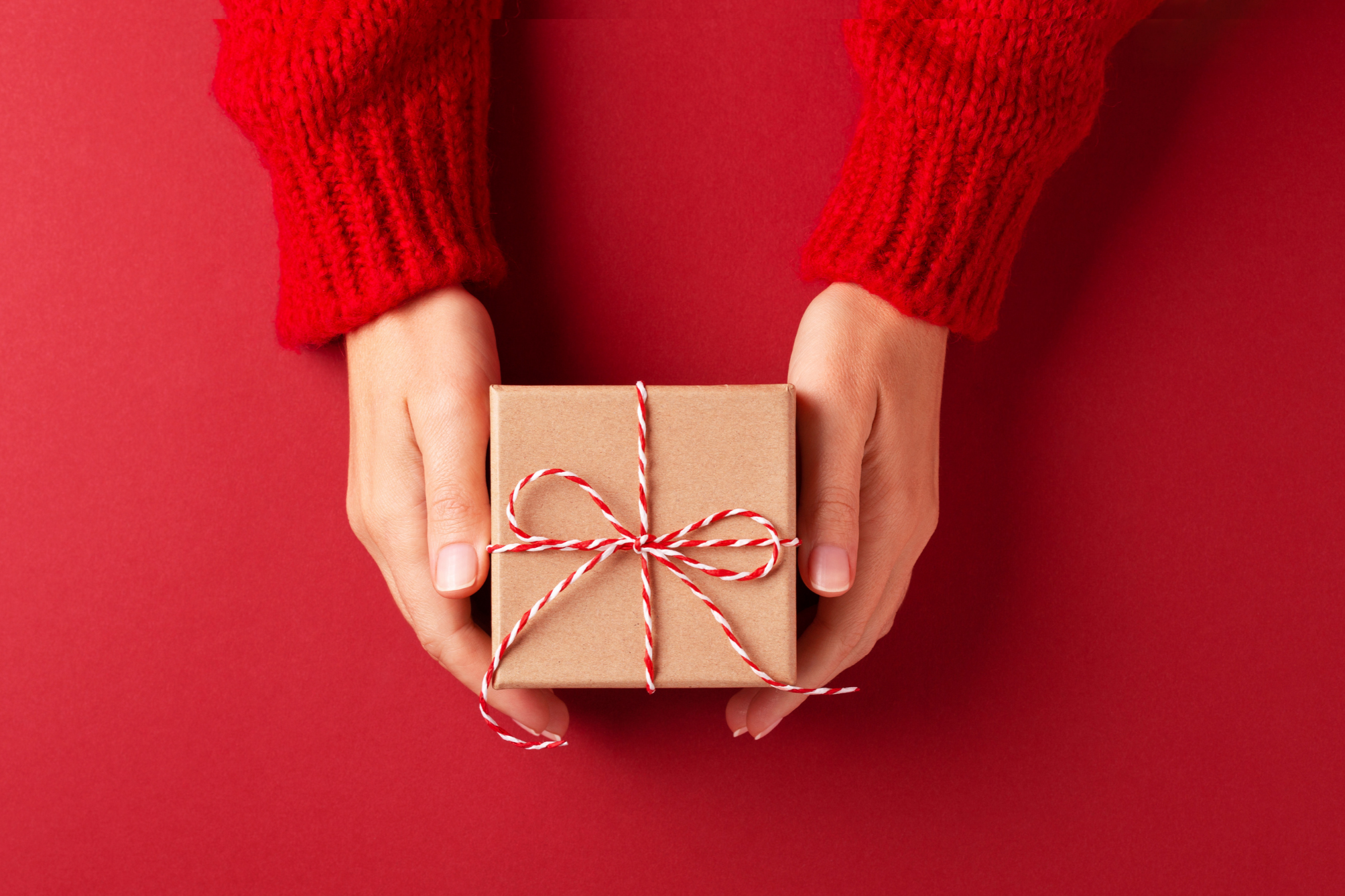 Person wearing red knitted sweater holding a gift wrapped in brown paper with red and white string, against a red background.