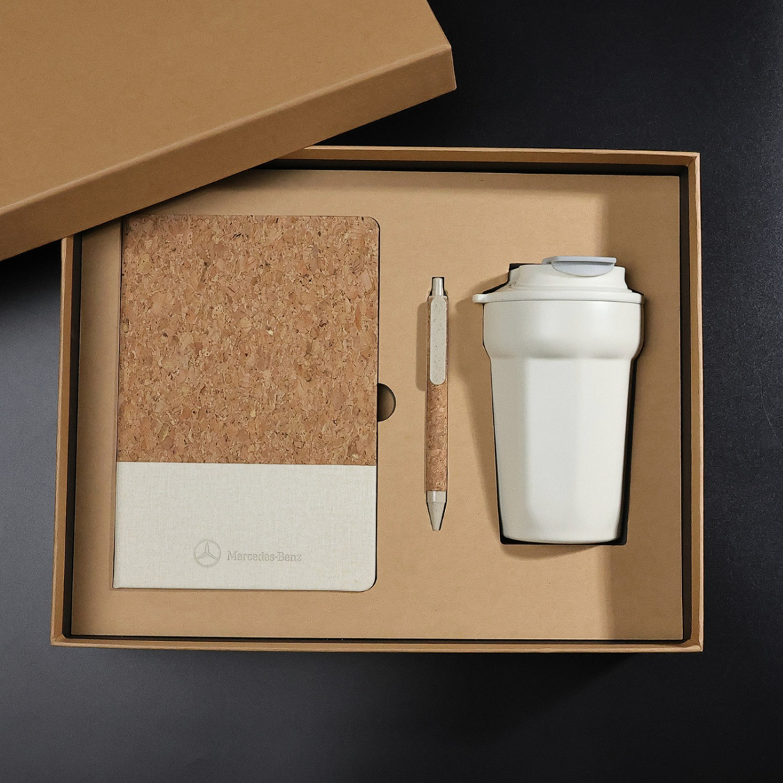 A gift box containing a notebook with cork cover, a pen, and a white reusable coffee cup with a lid.