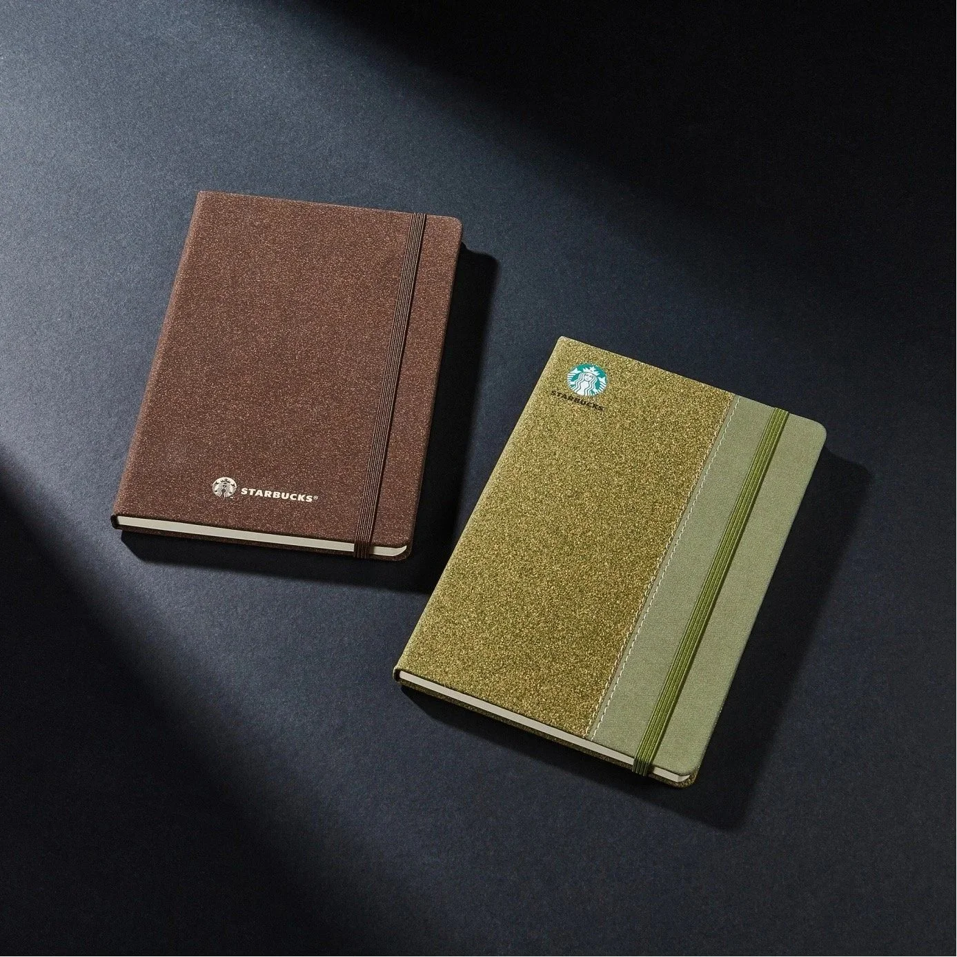Two Starbucks notebooks on a dark surface, one brown with a logo and elastic band, and the other green with a logo and elastic band, with shadows cast across the surface.