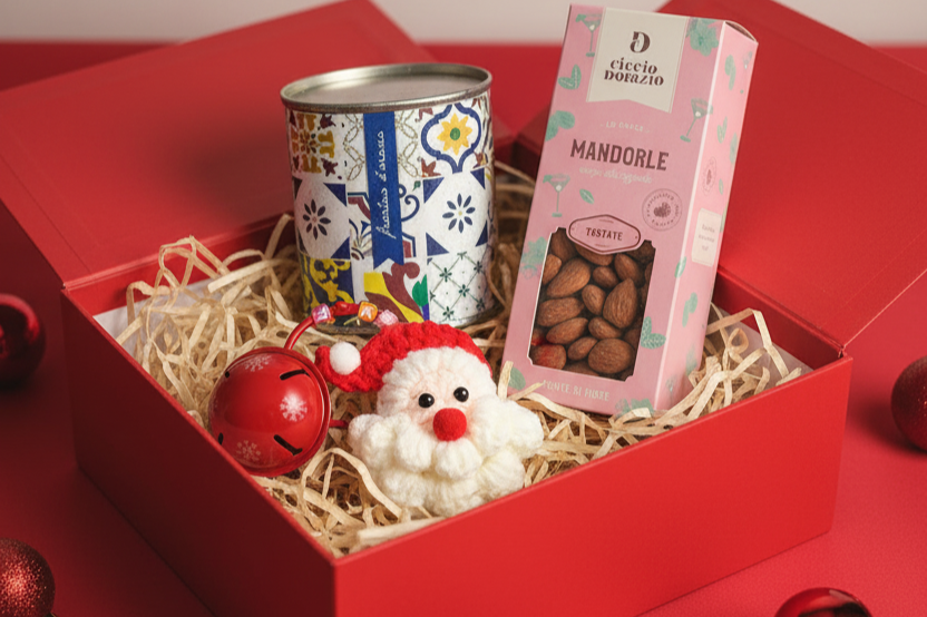 Christmas gift box containing a colorful patterned tin, a box of almond candies, a red Christmas bell, and a crocheted Santa Claus figurine, all on shredded paper inside a red box.