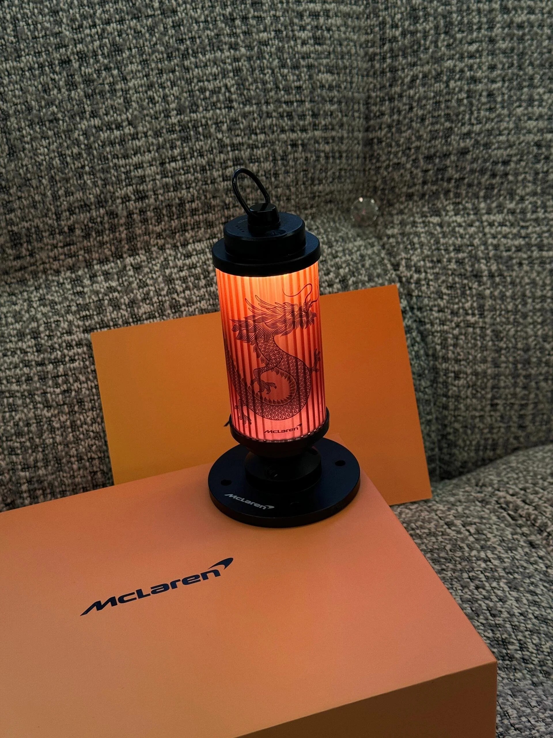 A McLaren branded lamp with a dragon illustration on the lampshade, placed on a McLaren box, on a textured gray fabric surface.