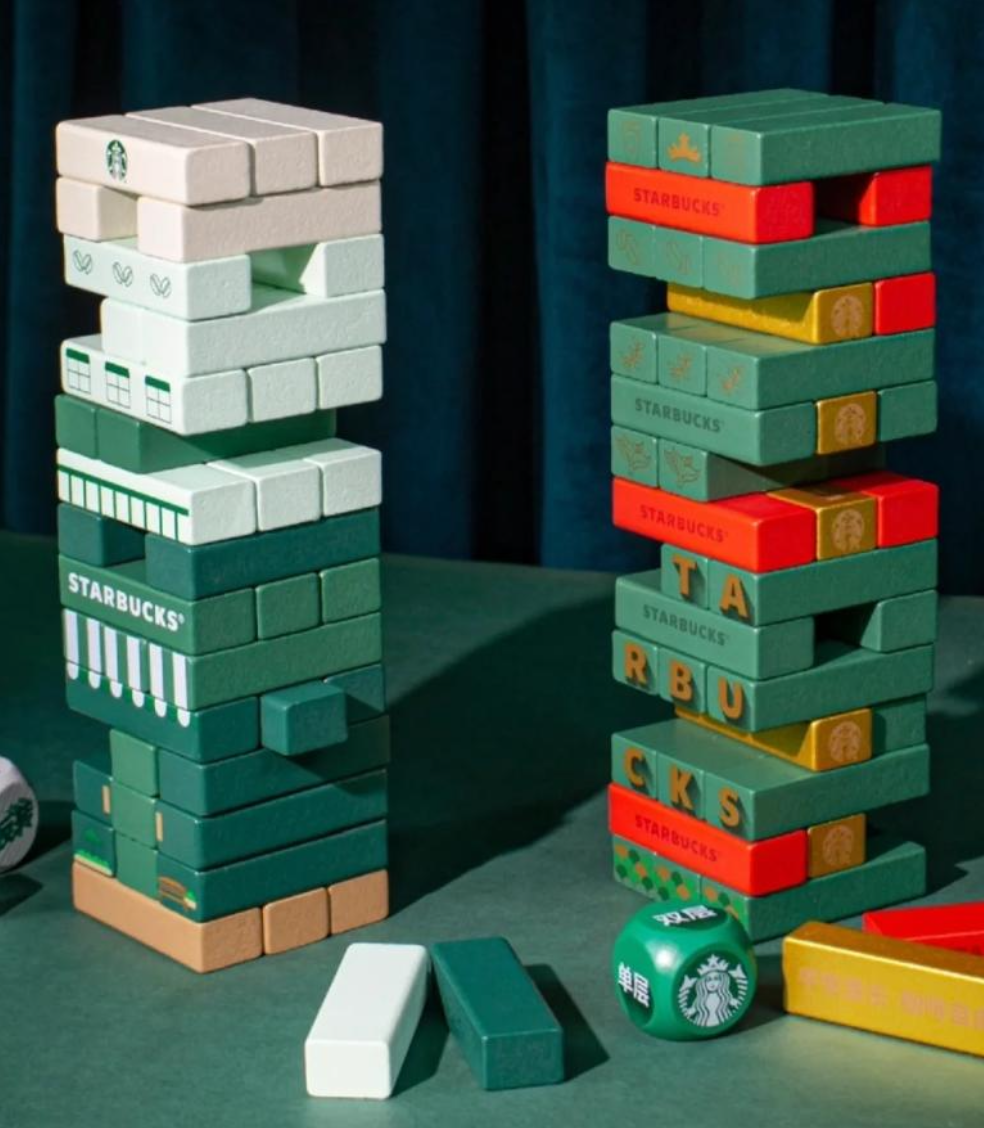 Two stacks of decorated domino tiles on a green table, with a Starbucks-themed die and domino pieces in front. The dominoes are themed with Starbucks logos, colors, and holiday designs.