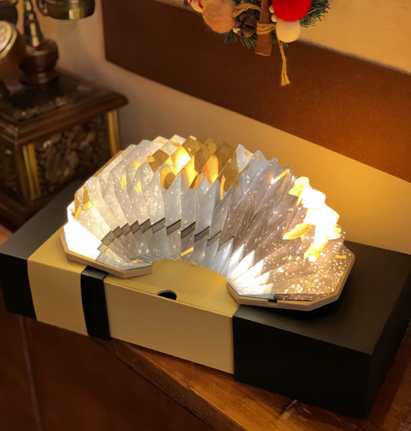 Decorative illuminated paper fan with star patterns on top of a black and white box, placed on a wooden surface, with a festive wreath hanging on the wall above.