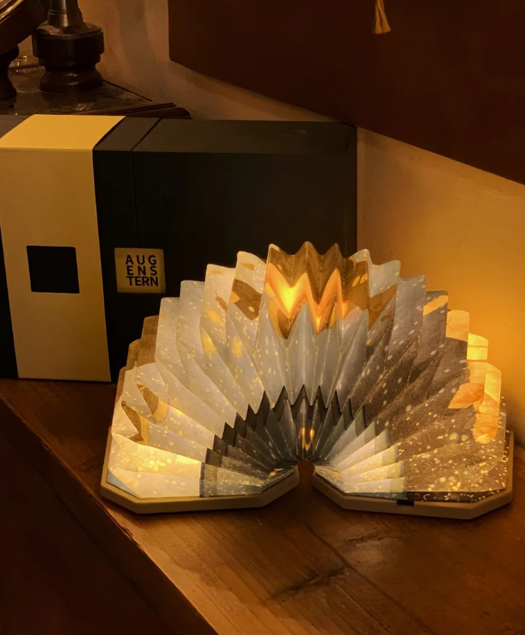 A decorative illuminated book-shaped sculpture with a starry night design, placed on a wooden surface next to a box labeled "AUG E N S T E R N."