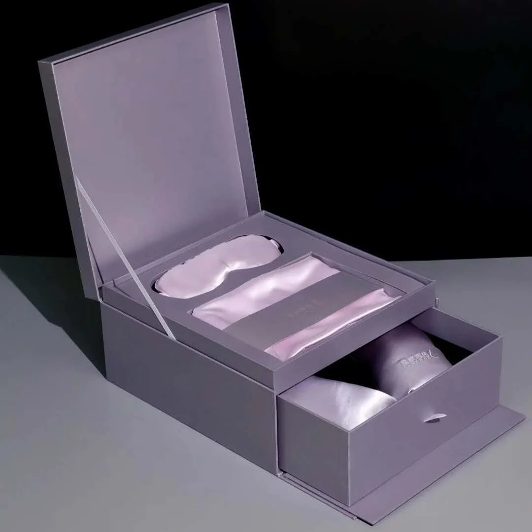 A silver gift box containing a satin sleep mask, a satin ribbon, and a small pouch inside a compartment, with a drawer at the front for storage.