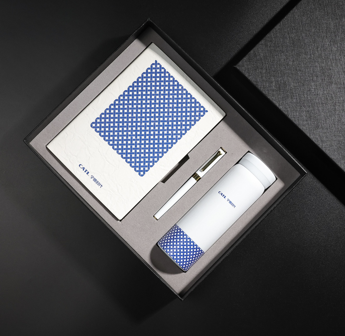 A gift set containing a white insulated tumbler with a blue geometric pattern at the base, a white notebook with a matching blue pattern on the cover, and a white pen with gold accents, all arranged inside a black box with gray foam padding.