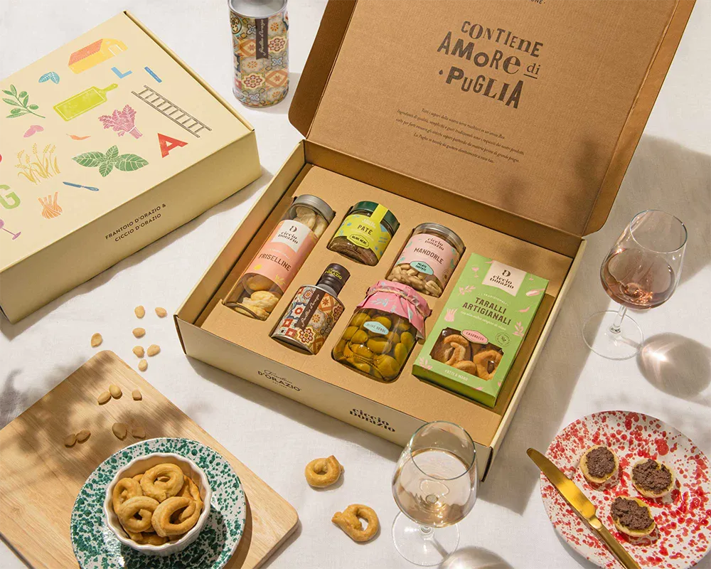 An open gift box with Italian food items, glasses of rosé wine, a cheese platter with crackers, and scattered snacks on a white table.