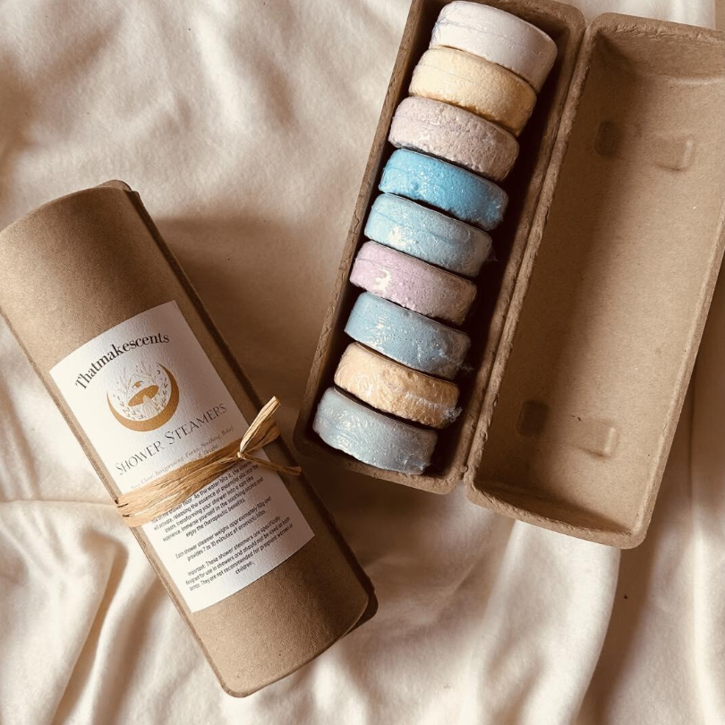 A cardboard tube labeled 'Shower Steamers' and a cardboard box containing pastel-colored, round bath bombs with a textured surface, arranged in a line inside the box, on a soft, cream-colored fabric surface.