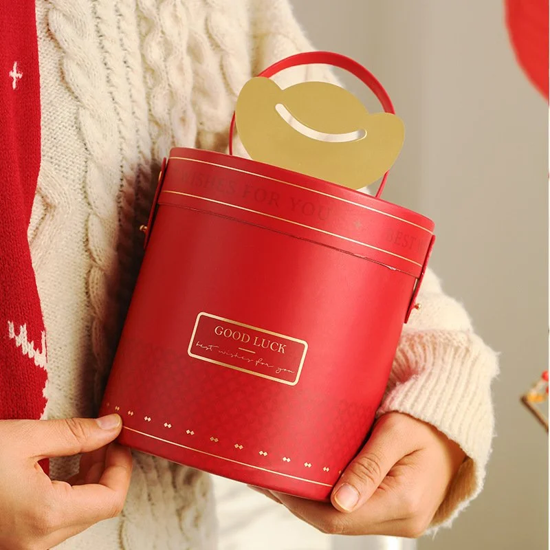 Person holding a red gift box with a gold smiley face paper inside, wearing a cream-colored sweater with a red and white knit scarf or blanket.