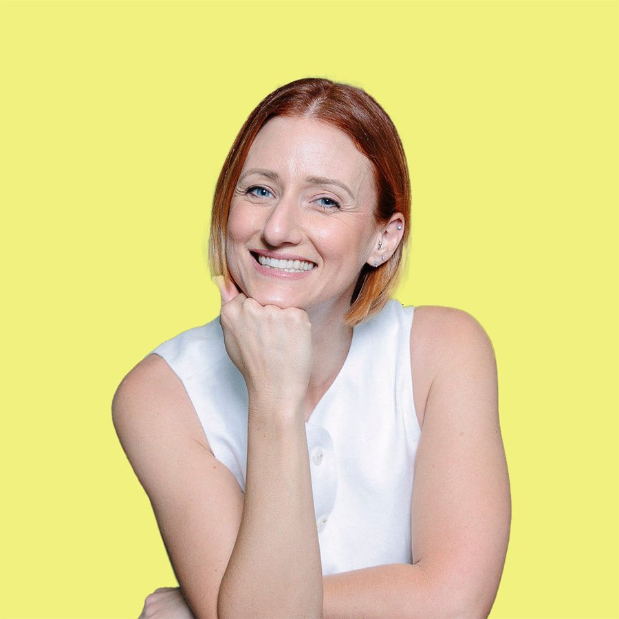 A young woman with red hair smiling and resting her chin on her hand, wearing a white sleeveless top, against a yellow background.