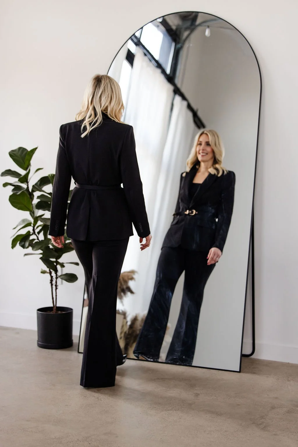 A woman in a black suit is looking at herself in a tall, oval mirror, smiling and adjusting her outfit. There is a green potted plant beside her and a window with white curtains behind.