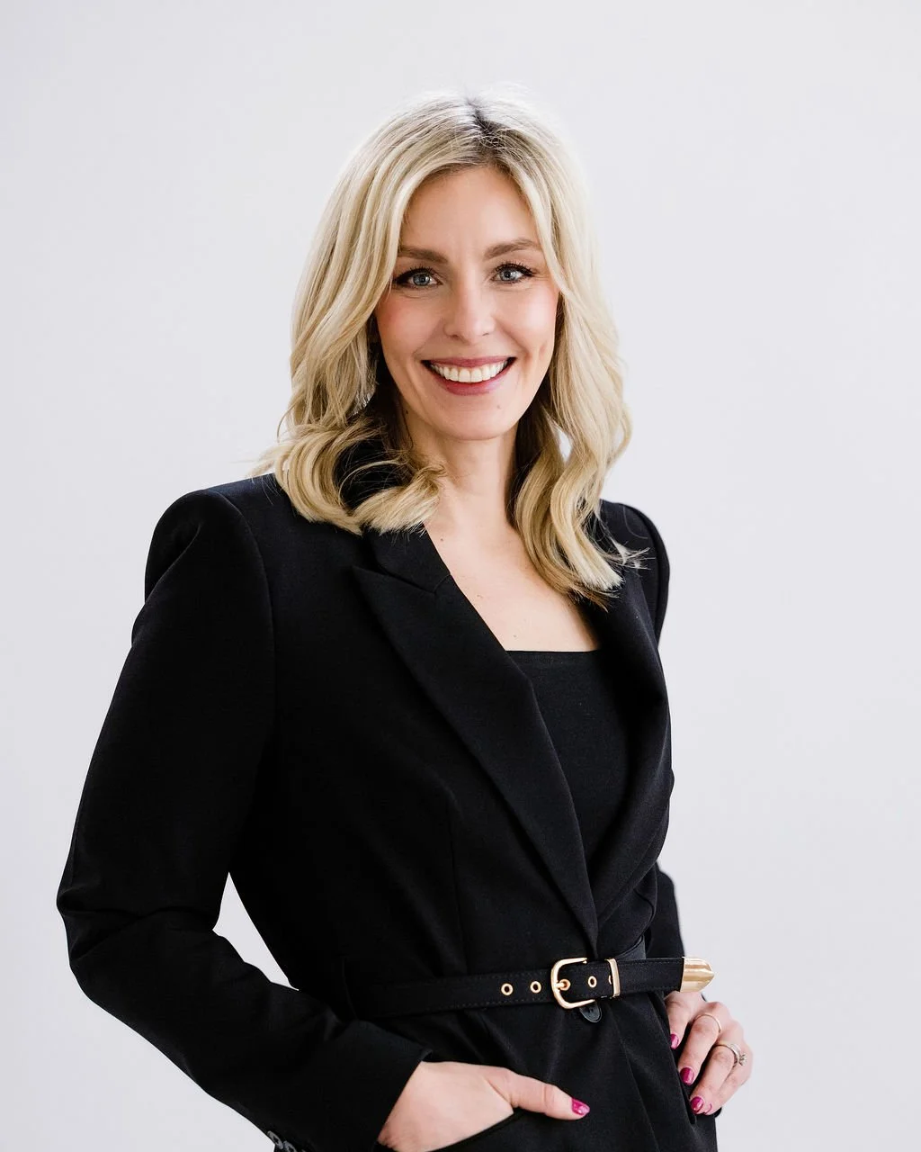 Professional woman with blonde wavy hair smiling, dressed in a black blazer and top, standing with hand on hip against a plain light gray background.