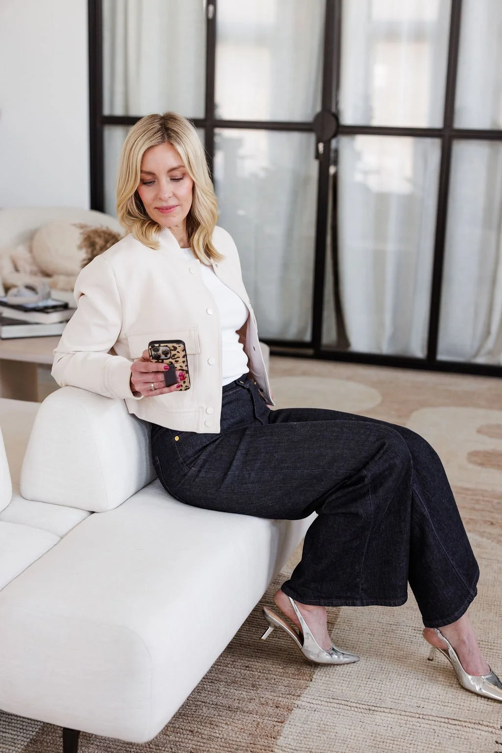 A woman sitting on a white sofa, looking at her phone with a leopard print case, in a modern living room with large windows and a beige rug.