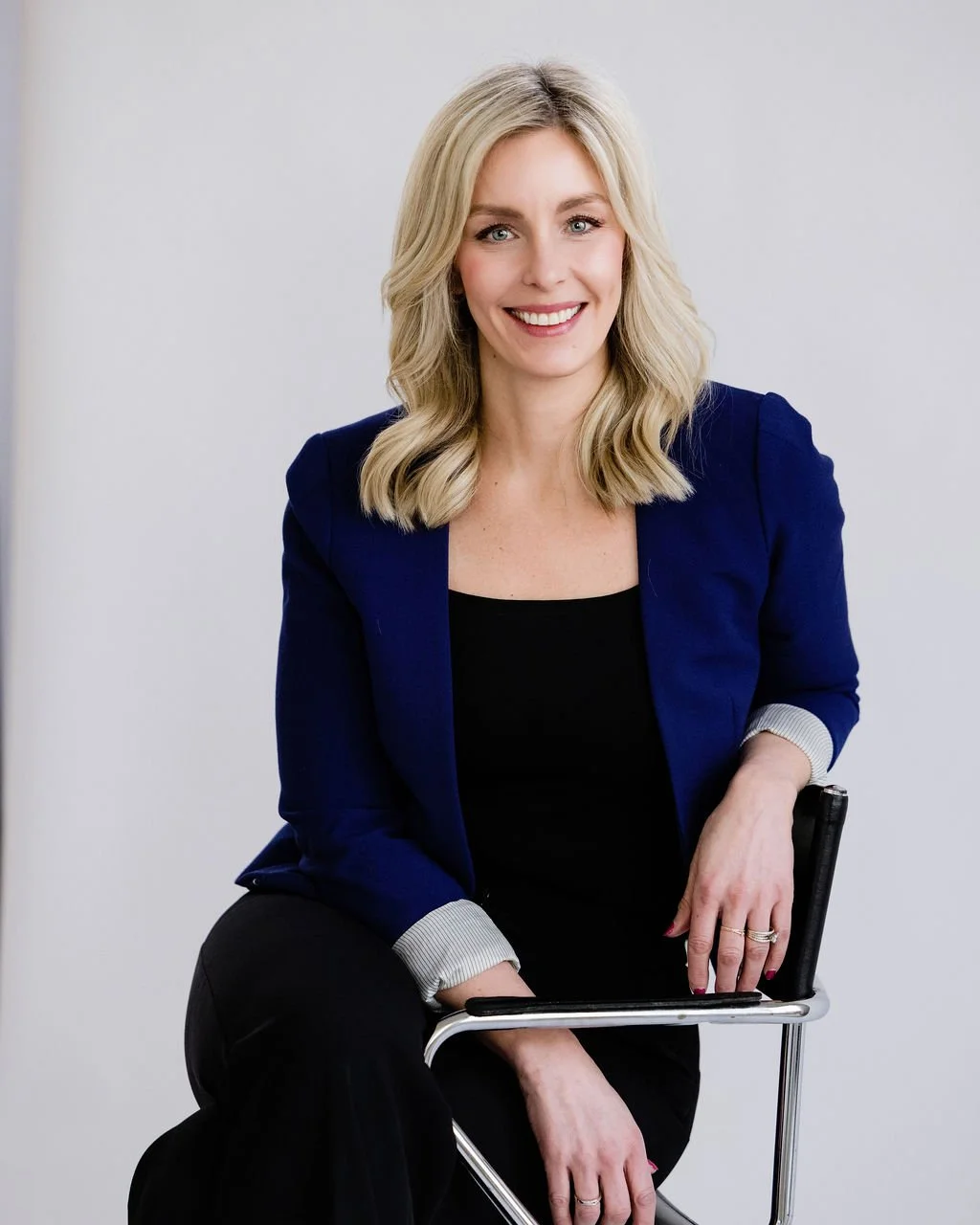 A woman with blonde hair, wearing a navy blazer and black top, smiling while sitting on a chair.