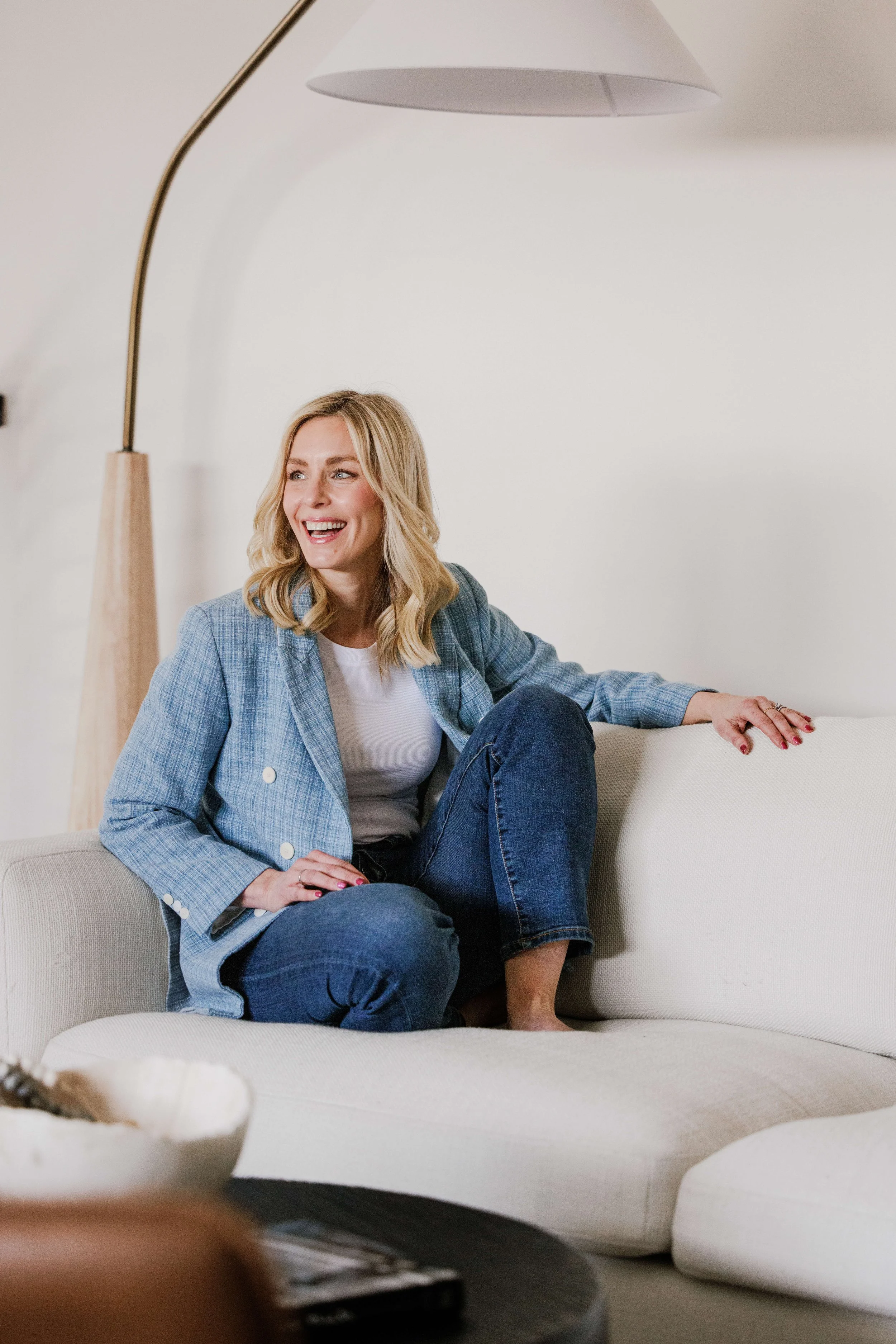 A smiling woman with blonde hair sitting on a white sofa, wearing a blue blazer and jeans, holding a relaxed pose in a modern, minimalistic living room.