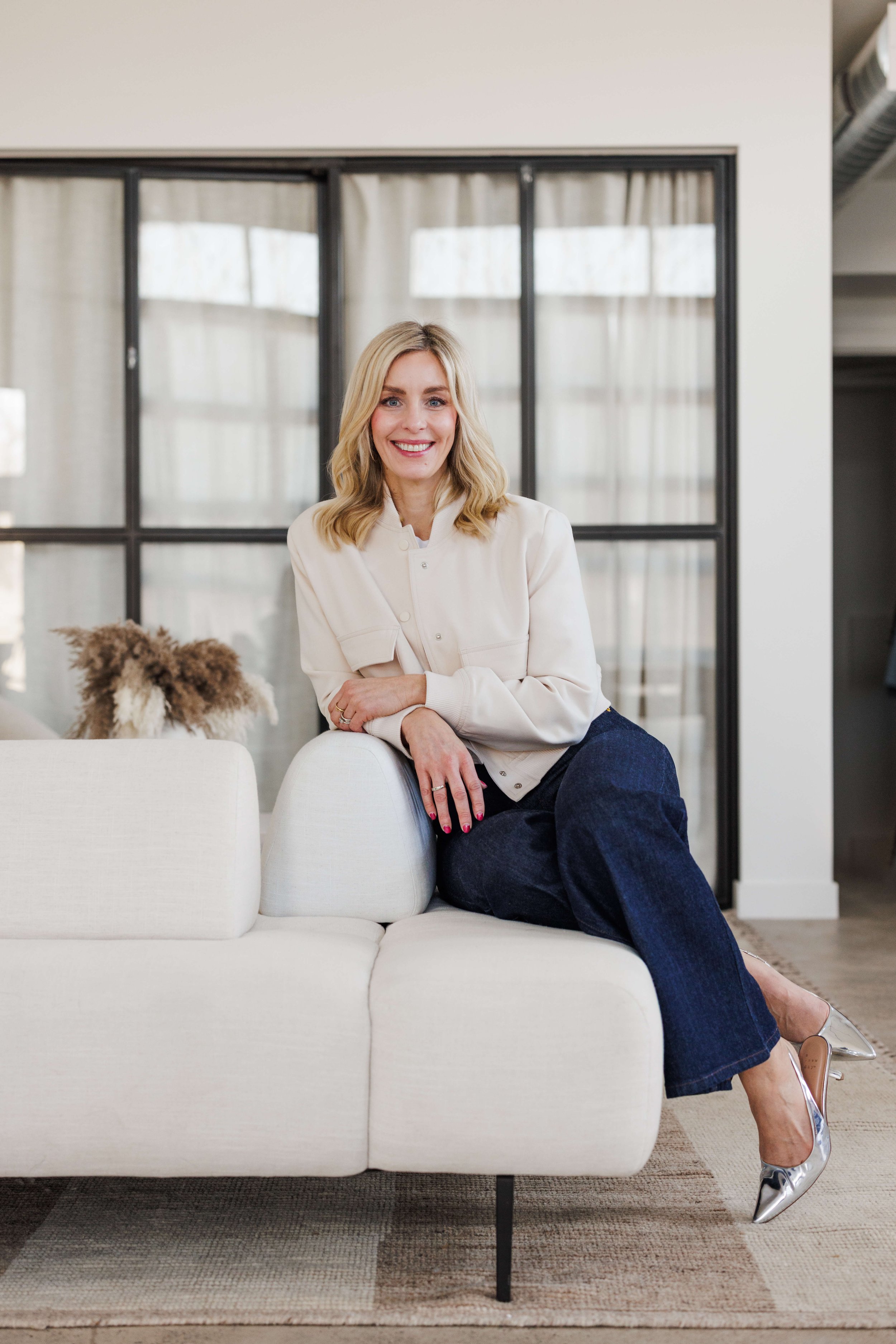 A woman with blonde hair sitting on the armrest of a white sofa, smiling, wearing a cream jacket, dark jeans, and metallic heels, in a bright, modern room with large windows and beige curtains.