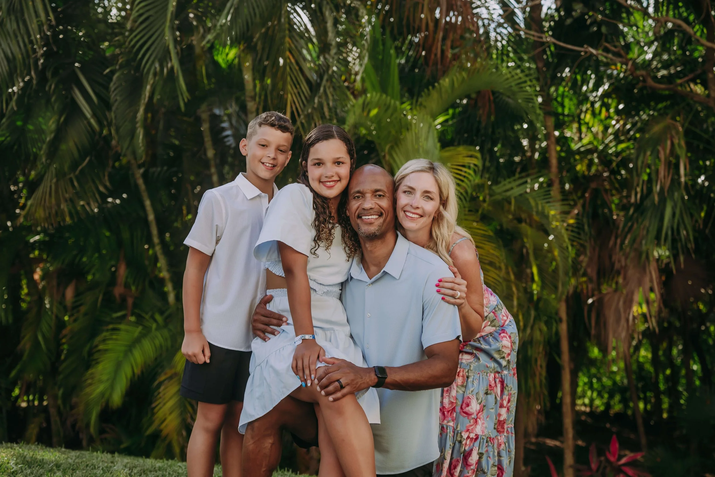 A happy multiracial family of five, including two children, a man, and two women, posing together outdoors among lush green tropical plants and trees, smiling at the camera.
