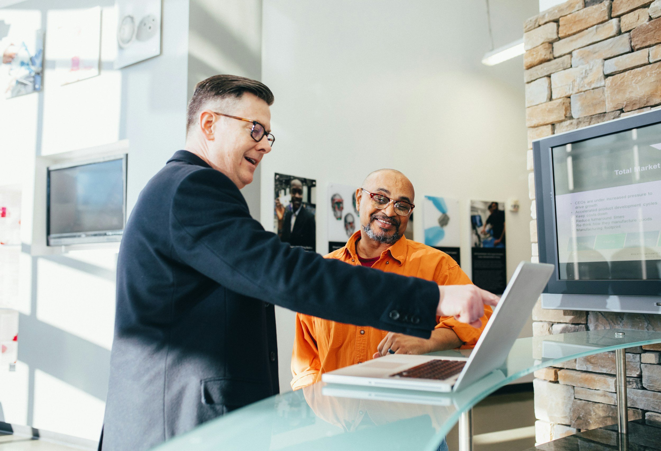 Two men are engaged in a discussion at a modern office reception desk, with one pointing at a laptop screen while the other looks on smiling.