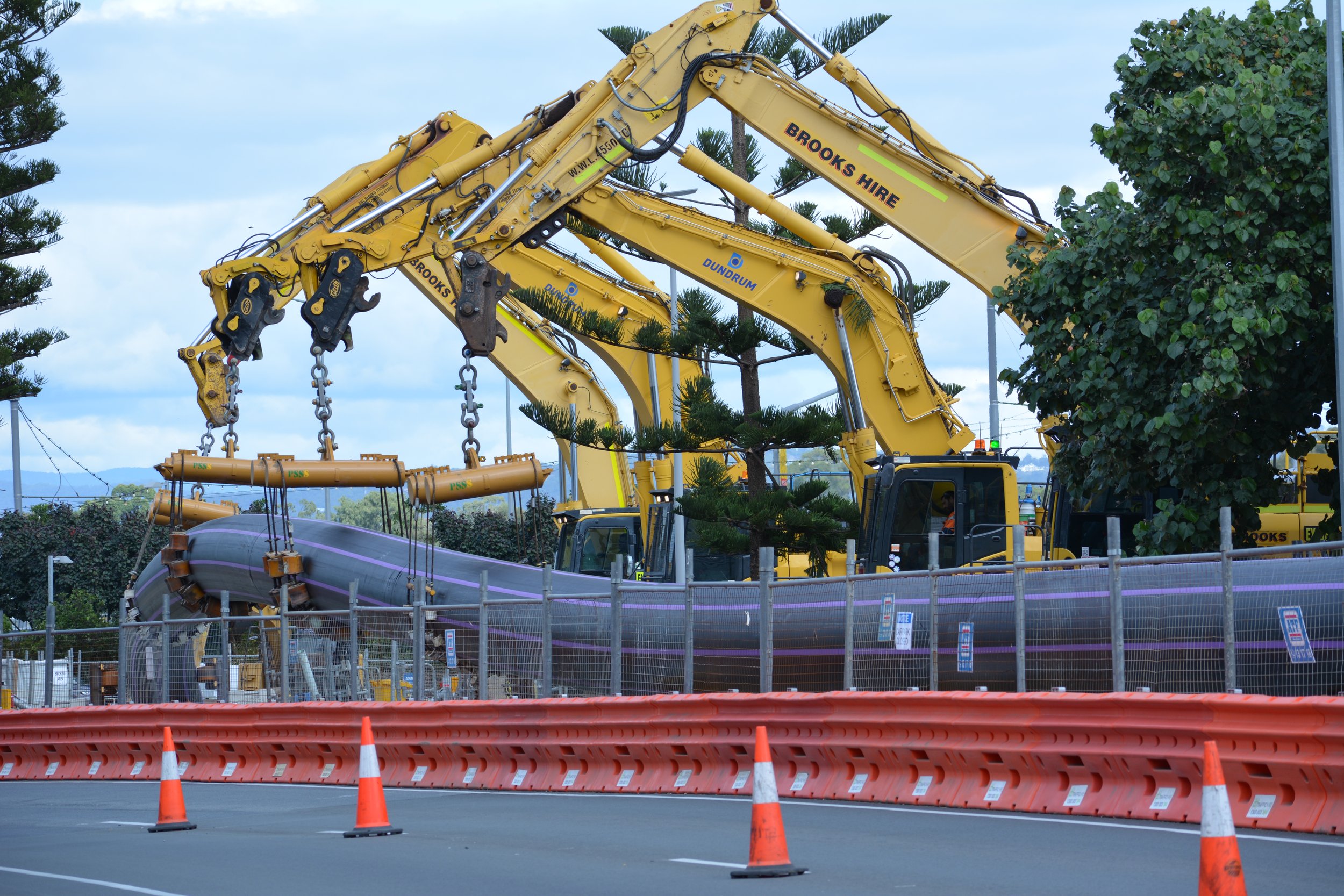Construction scene with multiple yellow excavators lifting a large pipeline, orange safety barriers, and traffic cones blocking the road, with trees and cloudy sky in the background.