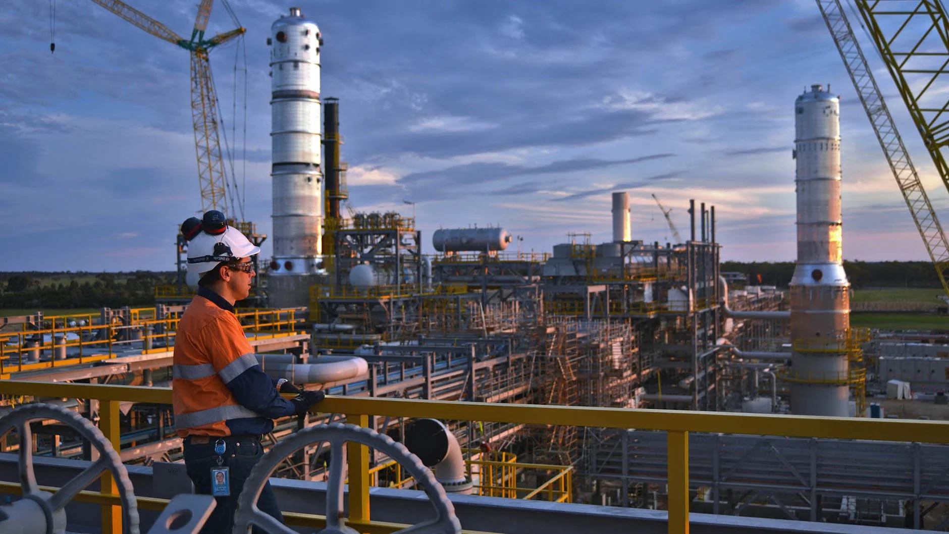 A worker in orange safety clothing stands on an industrial platform overlooking a large refinery with tall towers and machinery at sunset.