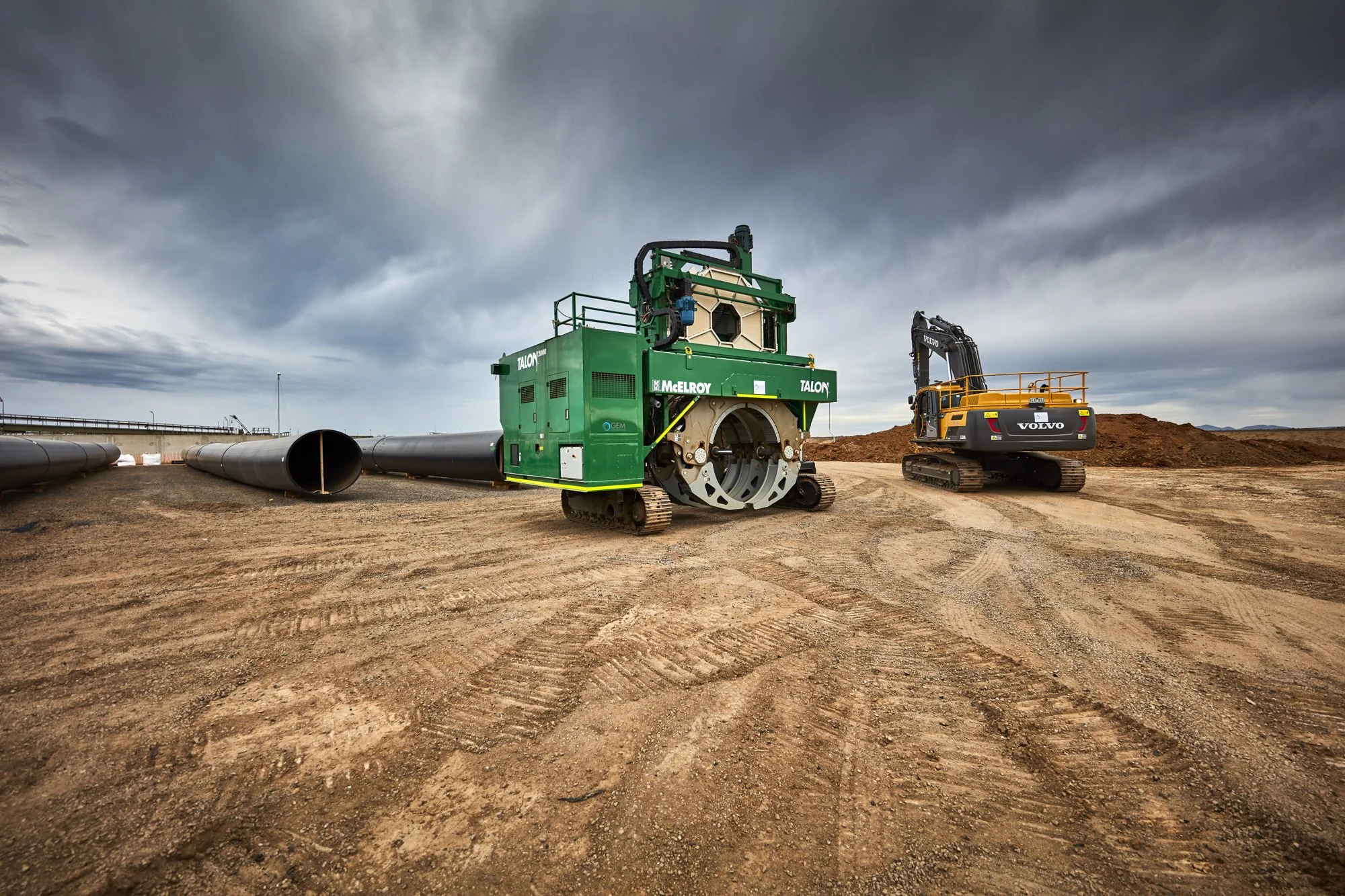 Construction site with large green and black pipe laying machine and yellow excavator on dirt, under cloudy sky.