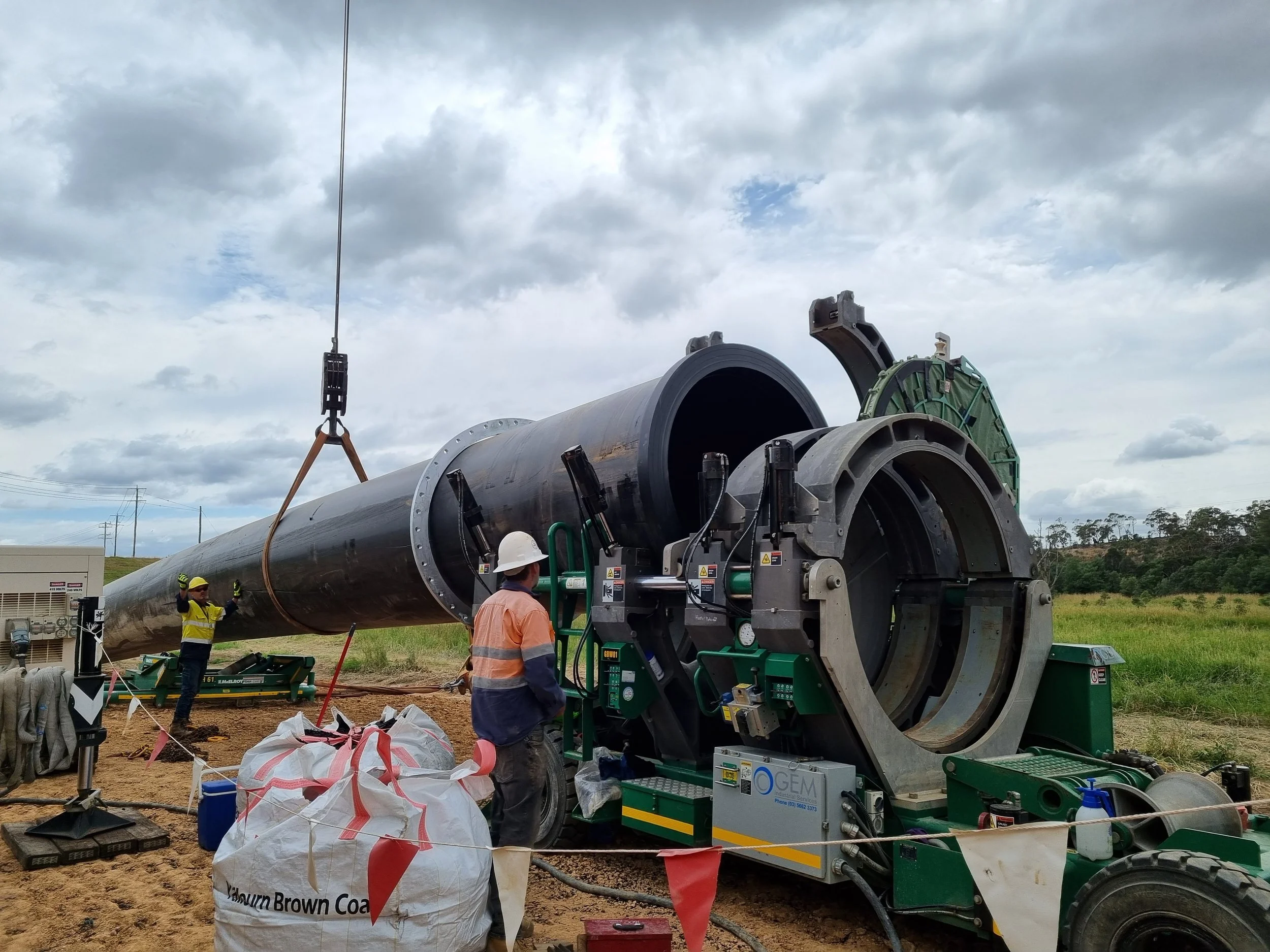 Construction workers install a large underground pipe using heavy machinery outdoors. The pipe is black and cylindrical, and the scene is set in an open field with cloudy skies.