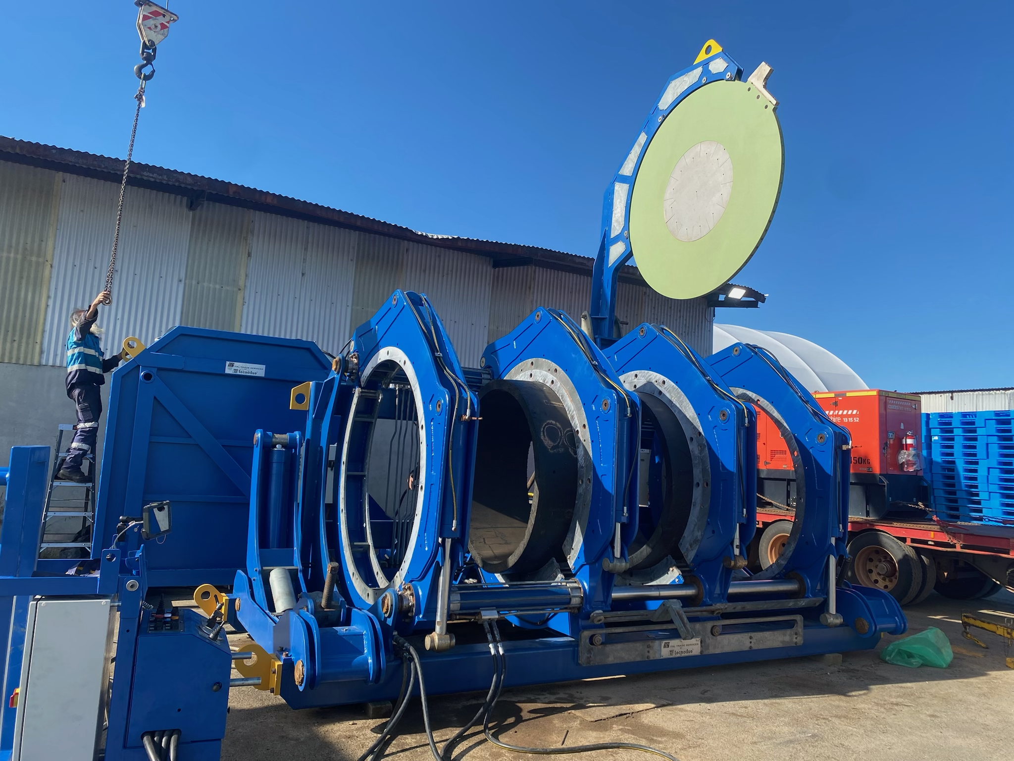 Large blue industrial machine with multiple cylindrical sections connected in a row, located outdoors under clear blue sky, with a person standing on a ladder working on the machine.