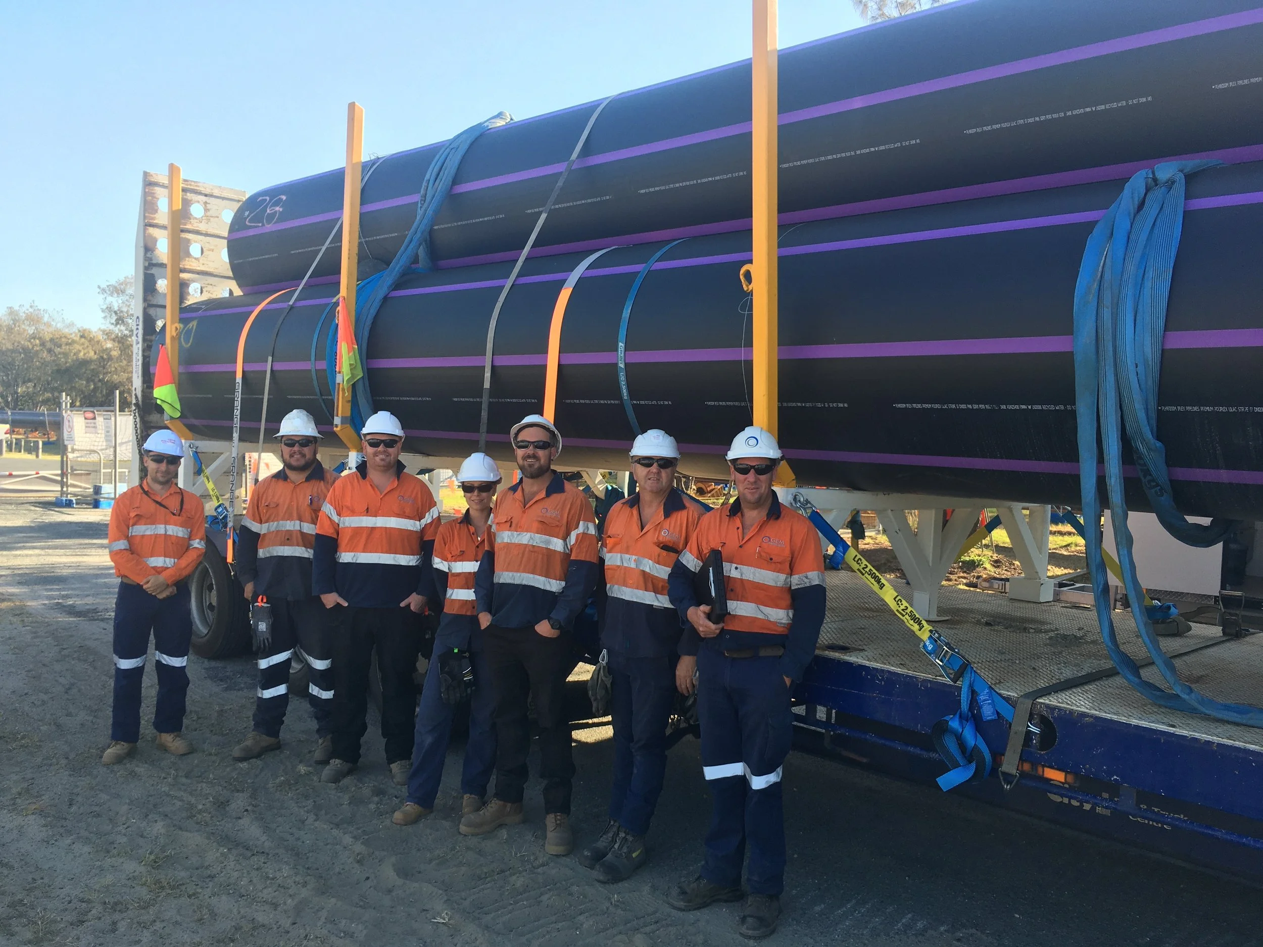Group of seven construction workers in safety gear standing next to a large aircraft wing or blade on a truck at a construction site