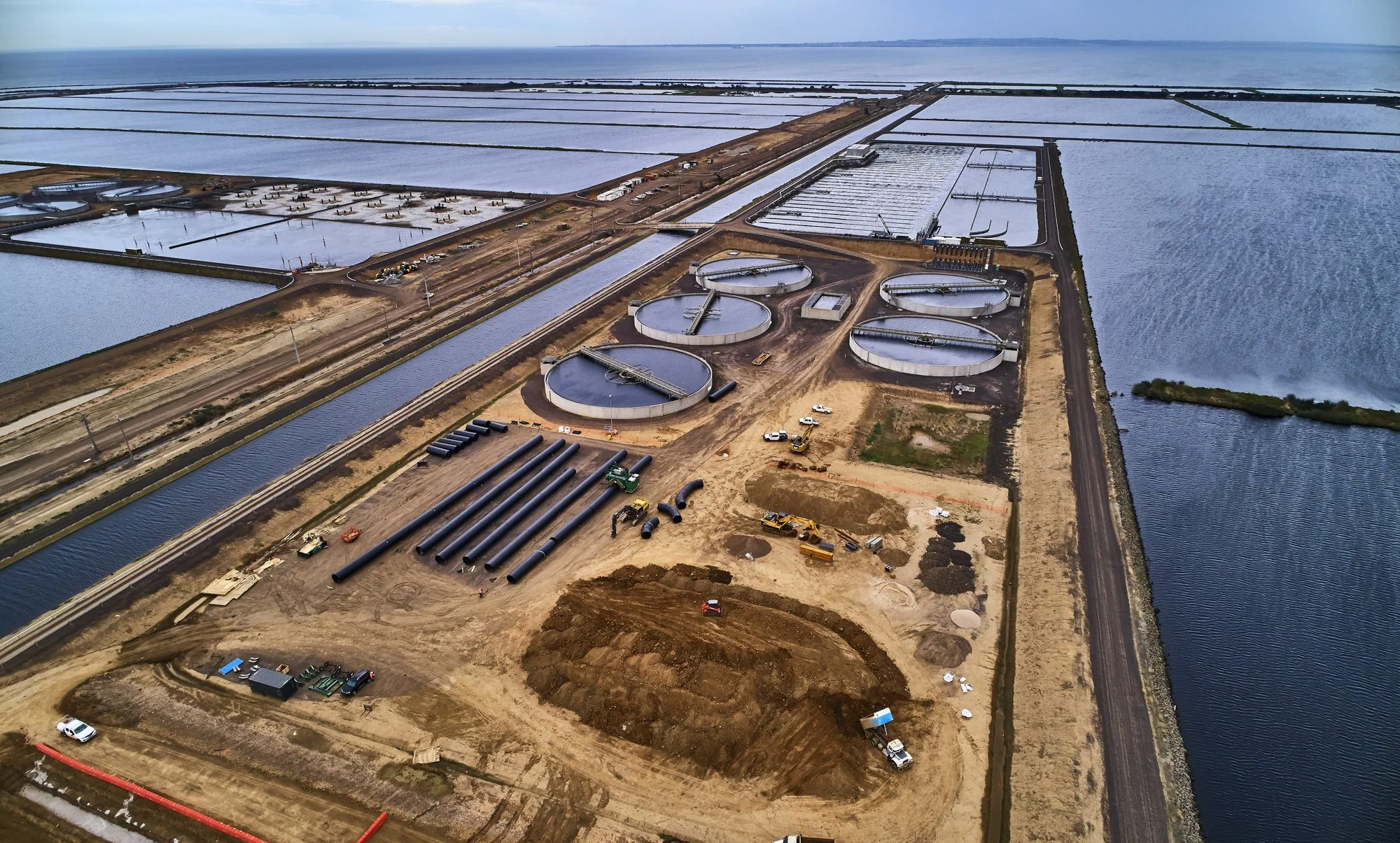Aerial view of a construction site near a body of water with large circular water treatment tanks, pipelines, construction vehicles, and ongoing infrastructure work.