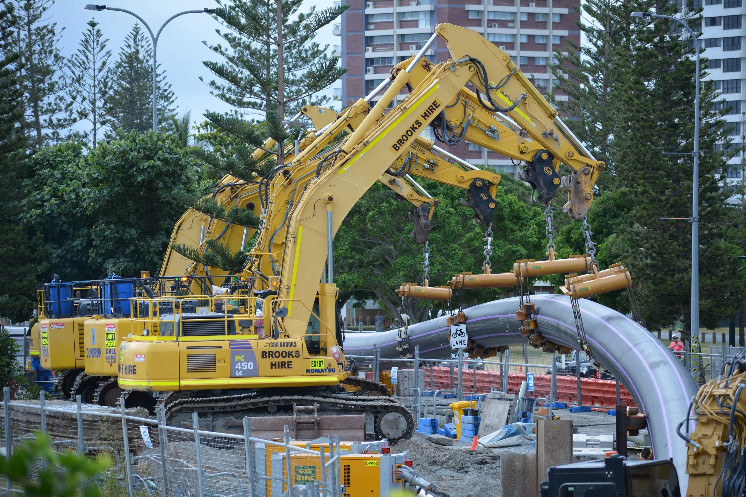 Construction site with large yellow excavator working on pipeline, surrounded by fencing and urban greenery.