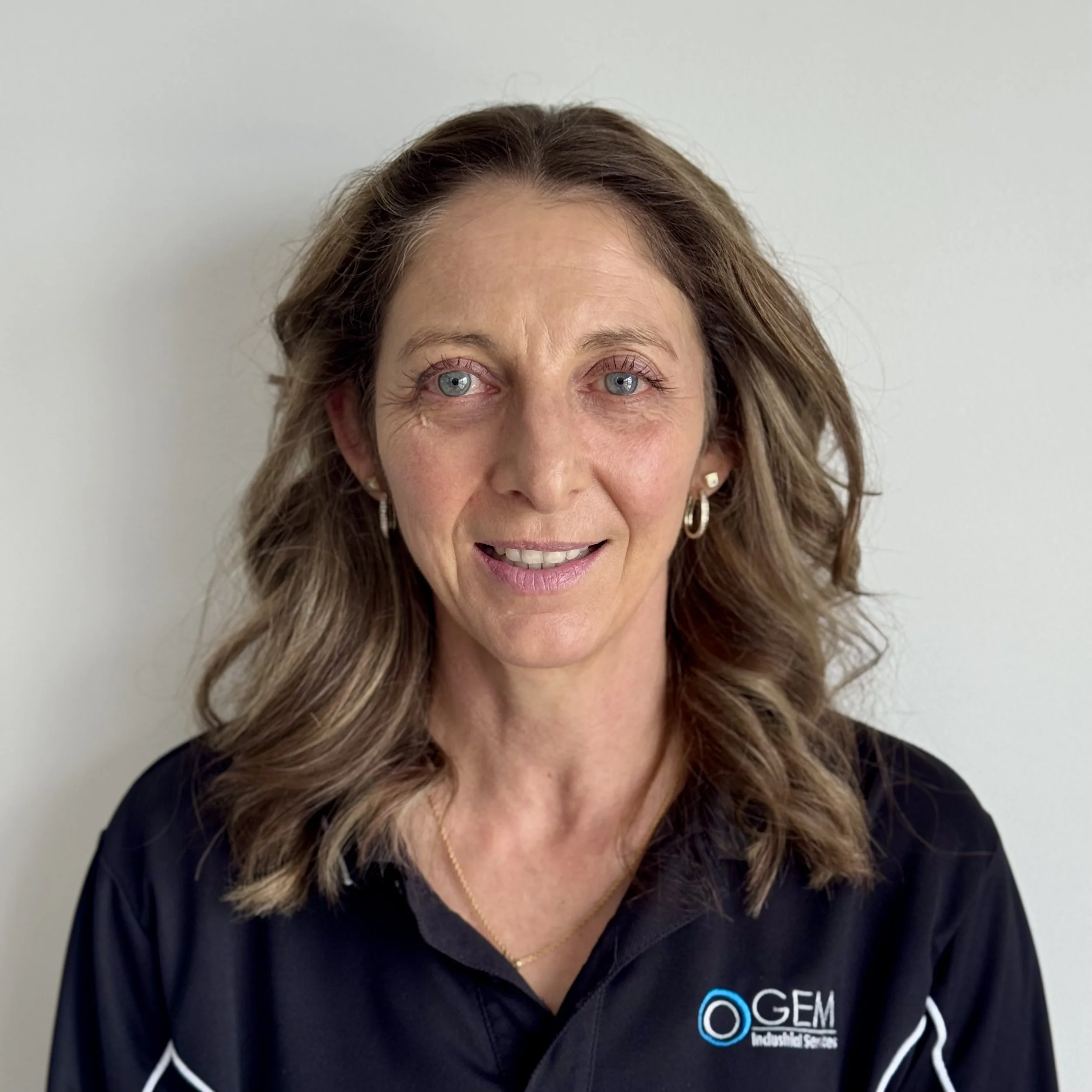 A woman with shoulder-length wavy brown hair and blue eyes, wearing a black collared shirt with a logo that reads 'GEM Industrial Services,' smiling slightly against a plain white wall.