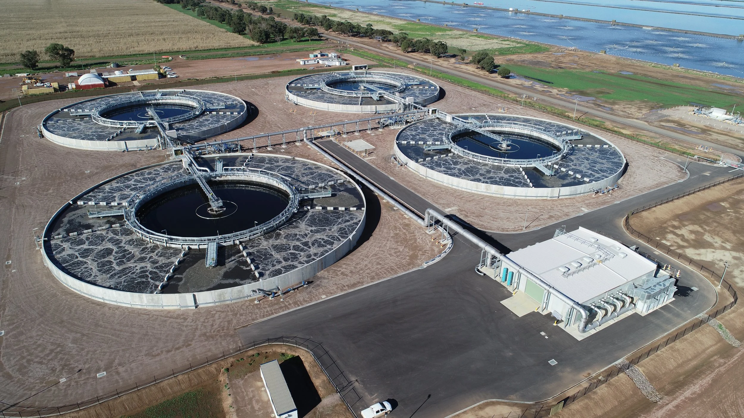 Aerial view of a water treatment facility with multiple circular clarifiers, pipelines, and a building, located near wetlands or salt ponds.