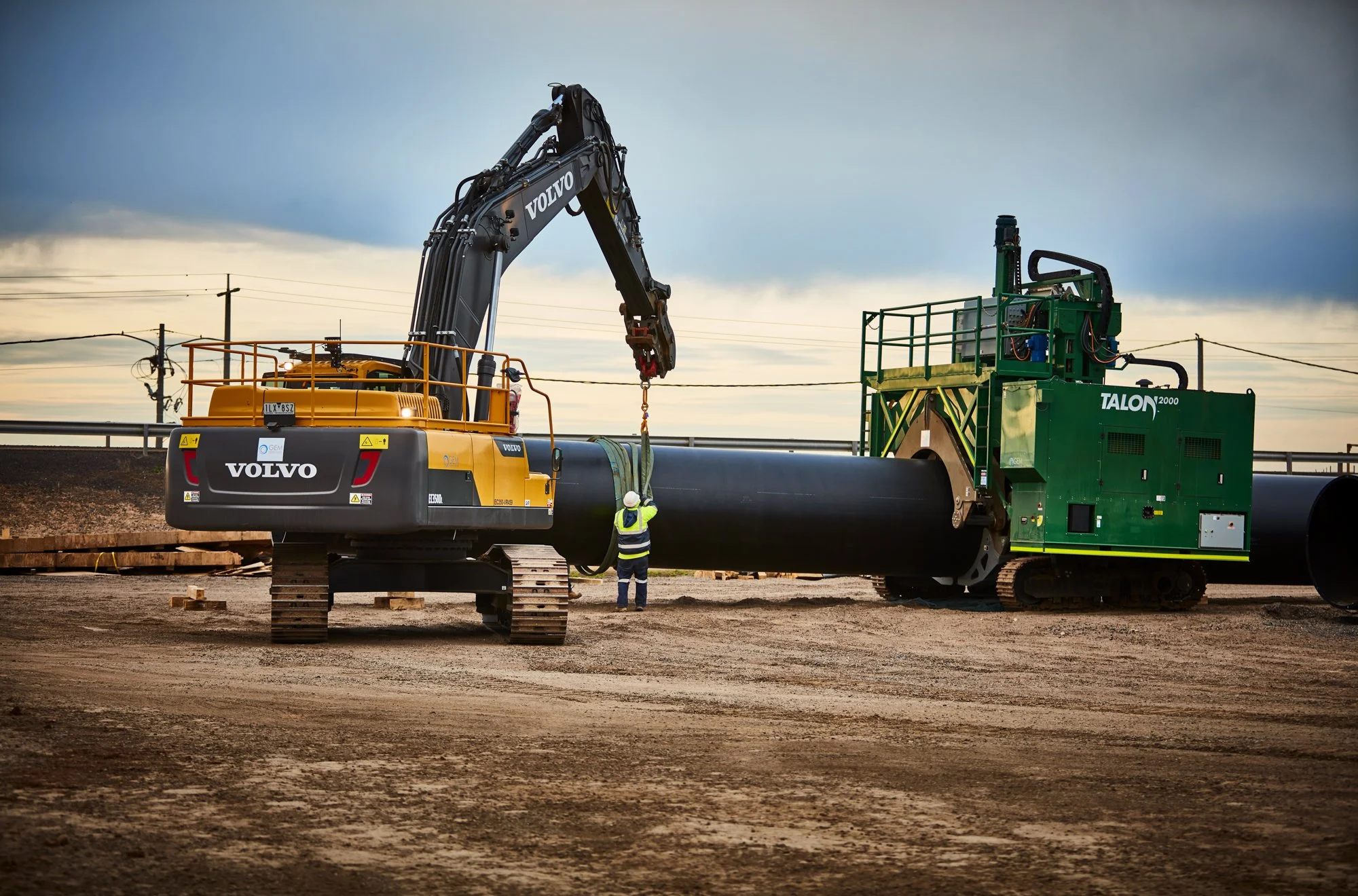 Construction workers are operating large industrial machinery on a dirt road in a construction site, with overhead power lines and a cloudy sky in the background.