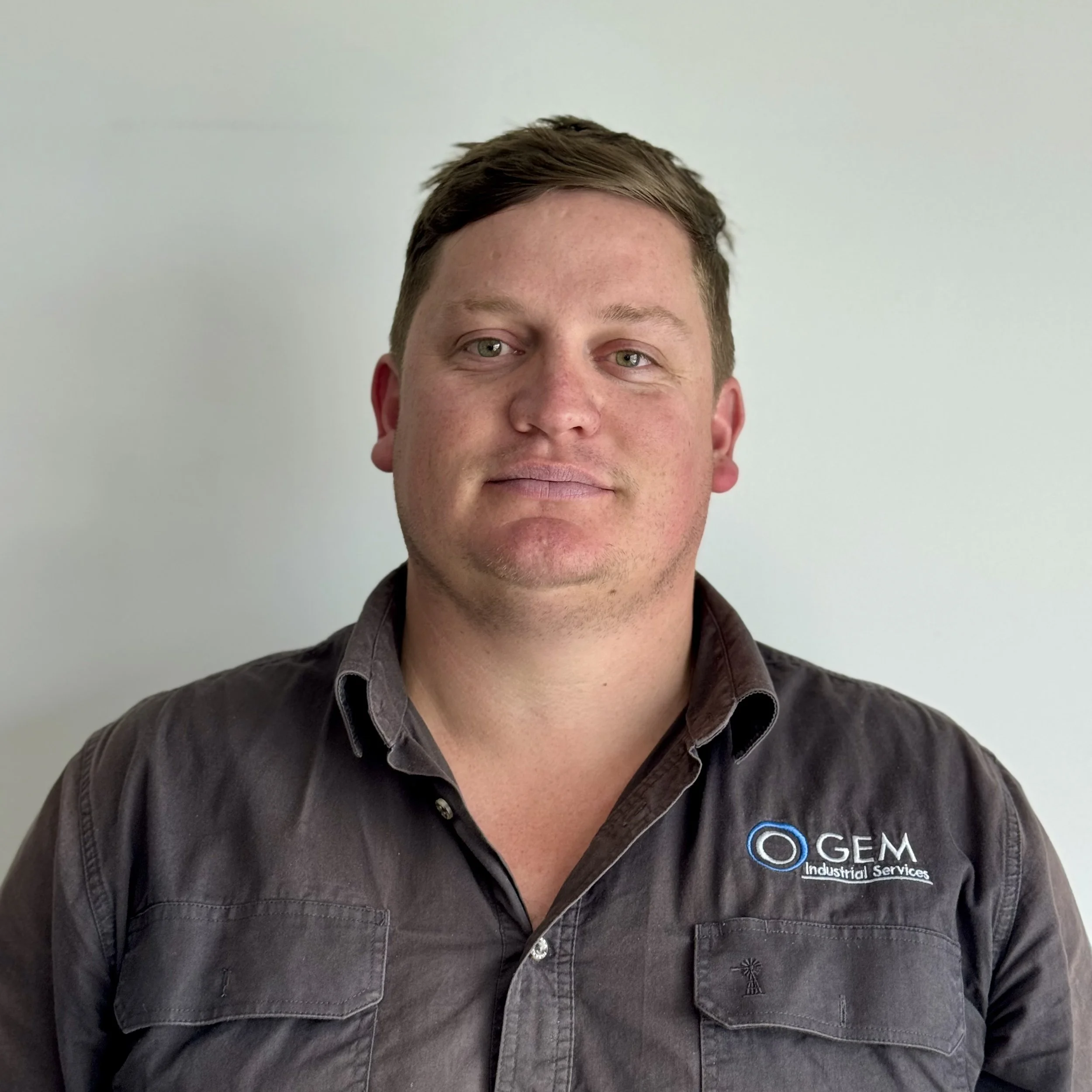 A man with short brown hair and fair skin wearing a dark gray button-up shirt with a logo on the chest that reads 'GEM Industrial Services'.