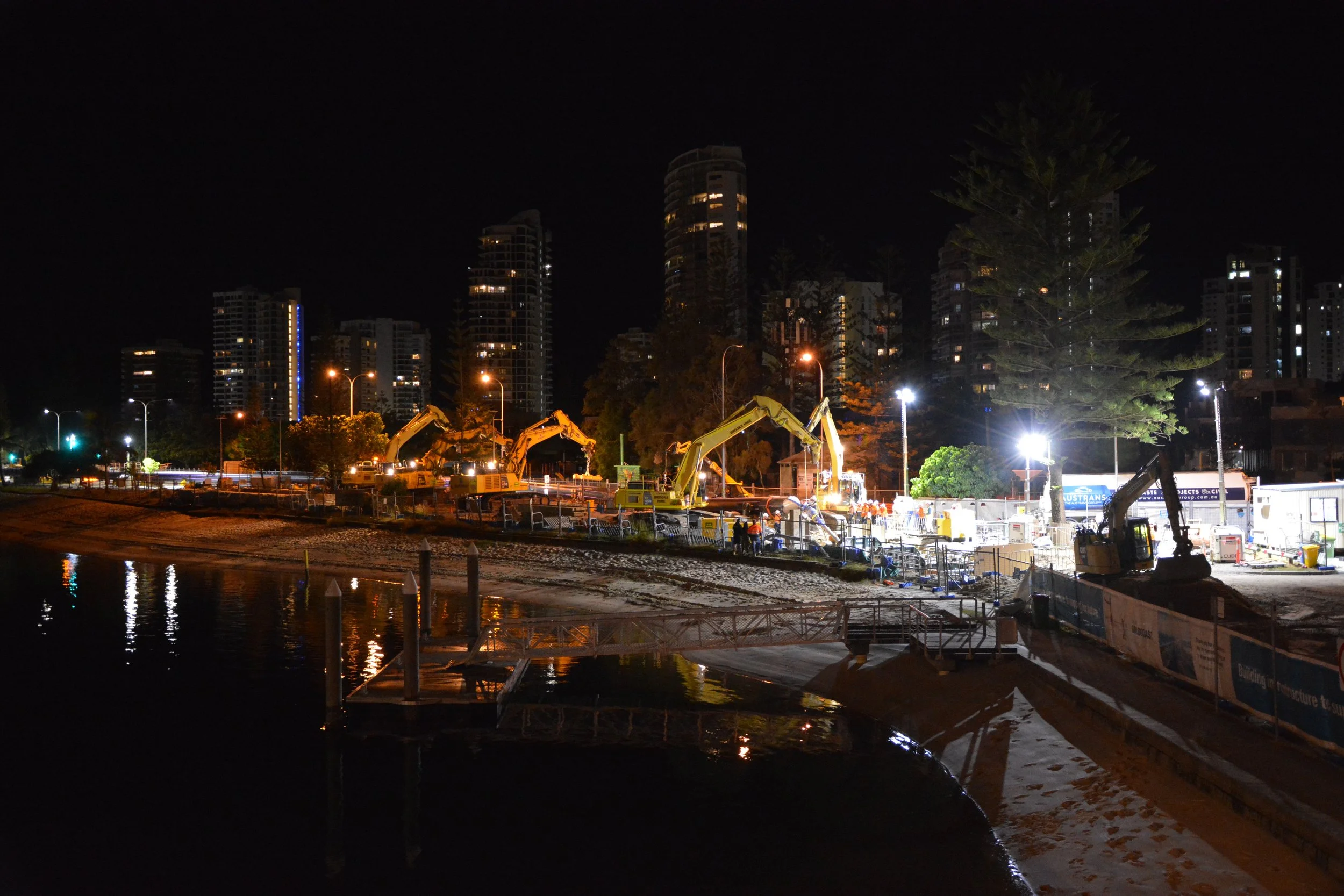 Nighttime construction site with excavators working near a water body, illuminated by bright lights, with tall buildings in the background.