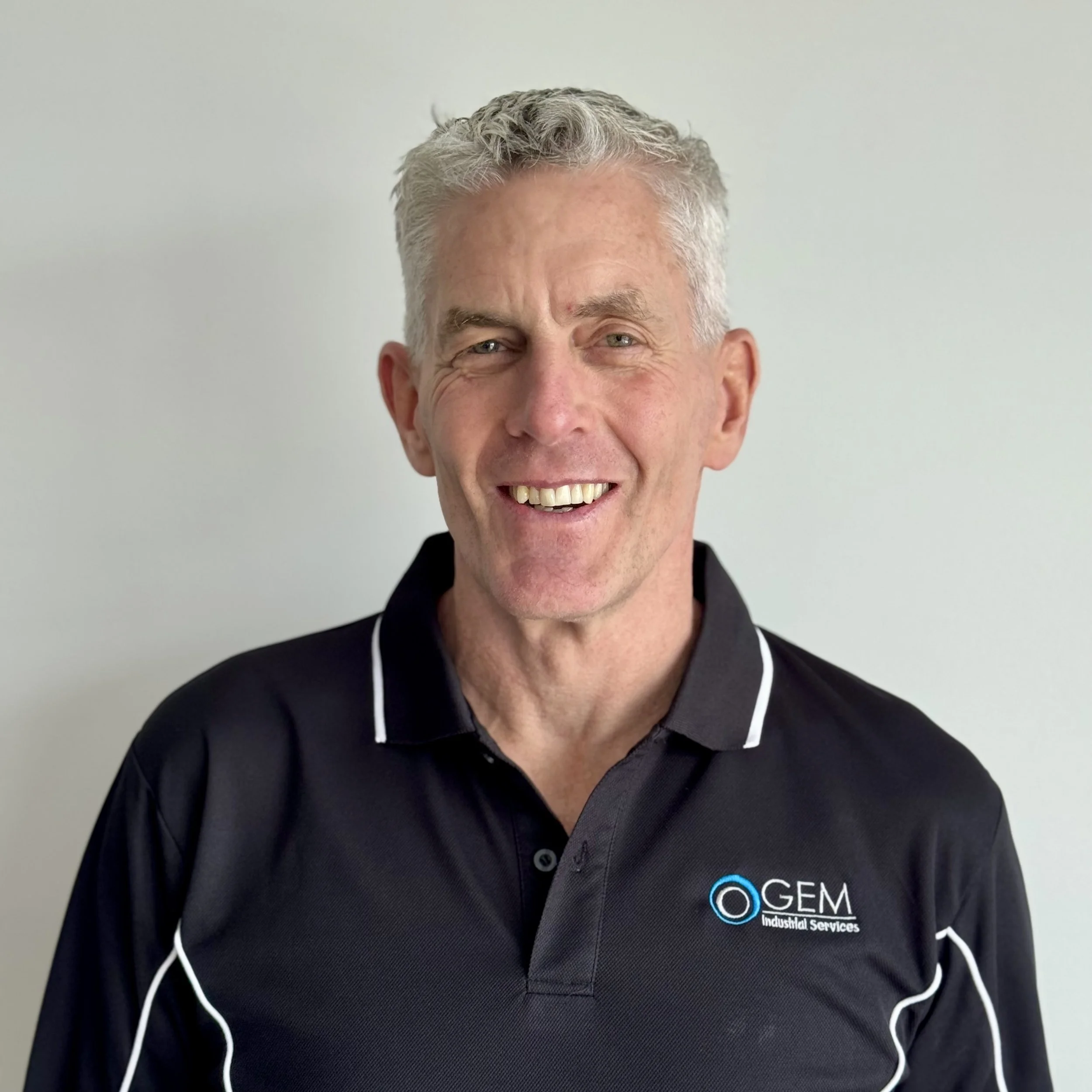 smiling man in black polo shirt with 'GEM Industrial Services' logo against plain white wall